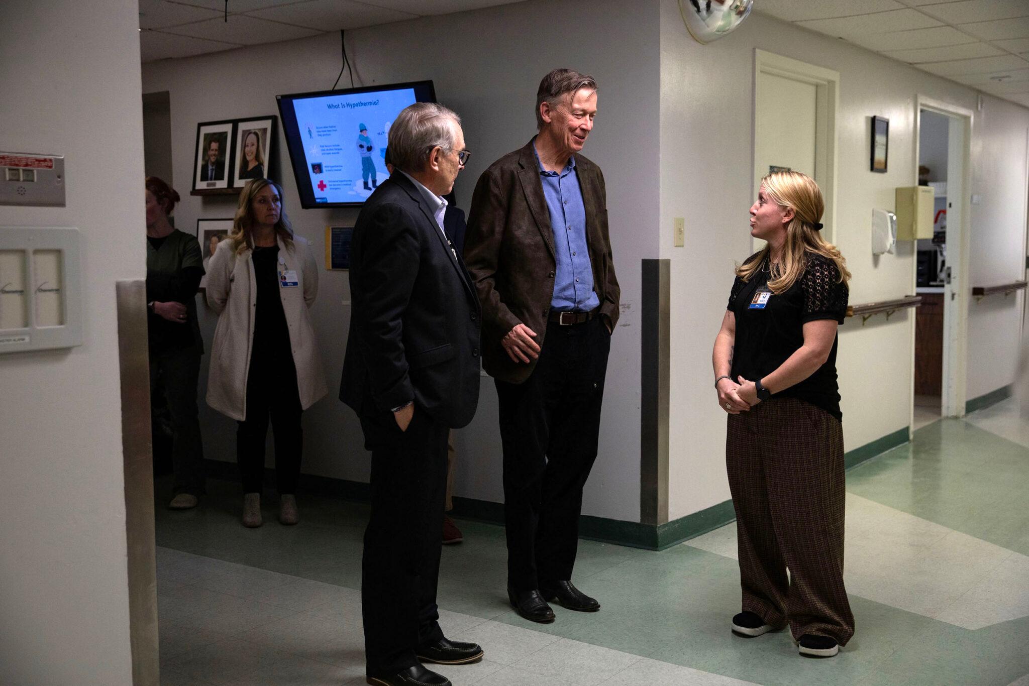 Two men and a woman chat in a hospital hallway with white walls and faded linoleum tile. A doctor in a white coat stands in shadow behind them.