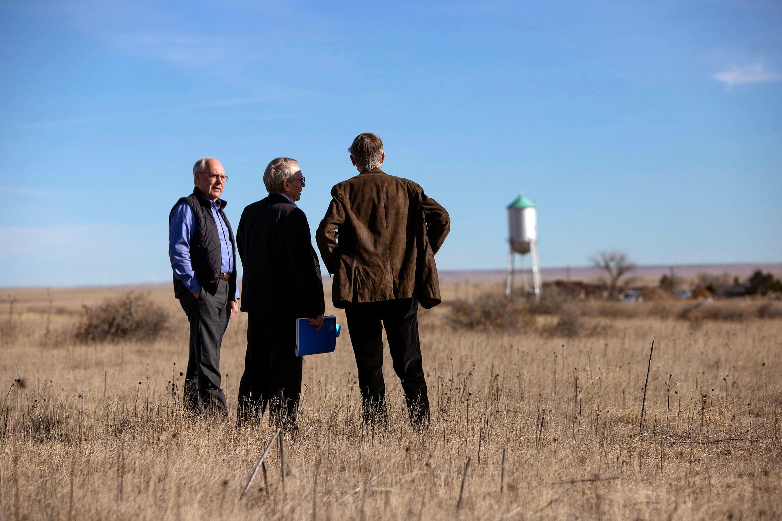 Three men stand in a dead winter field, staring into the distance which is empty, save for a water tower on the horizon.