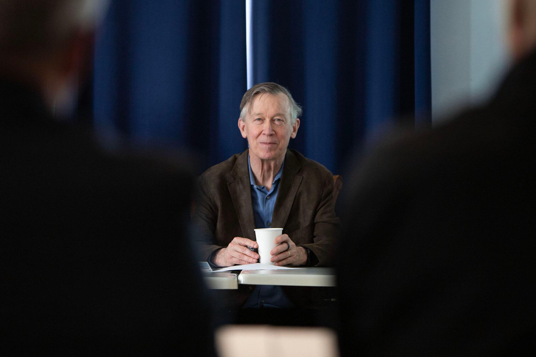 An older man in a brown jacket can be seen through the shoulders of two people sitting between him and the camera, smiling slightly right into the lens.