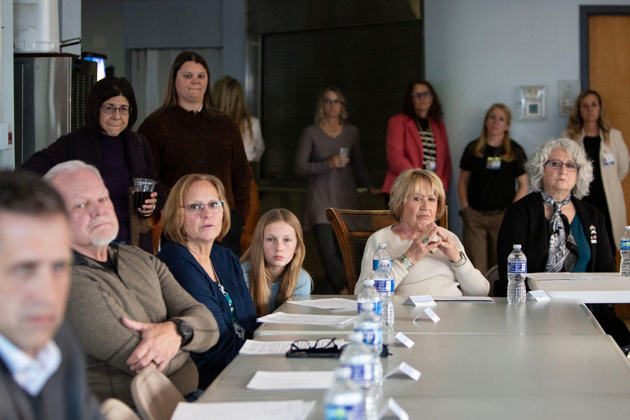 A group of people sit around a table, looking sternly as they listen to someone speaking off screen.
