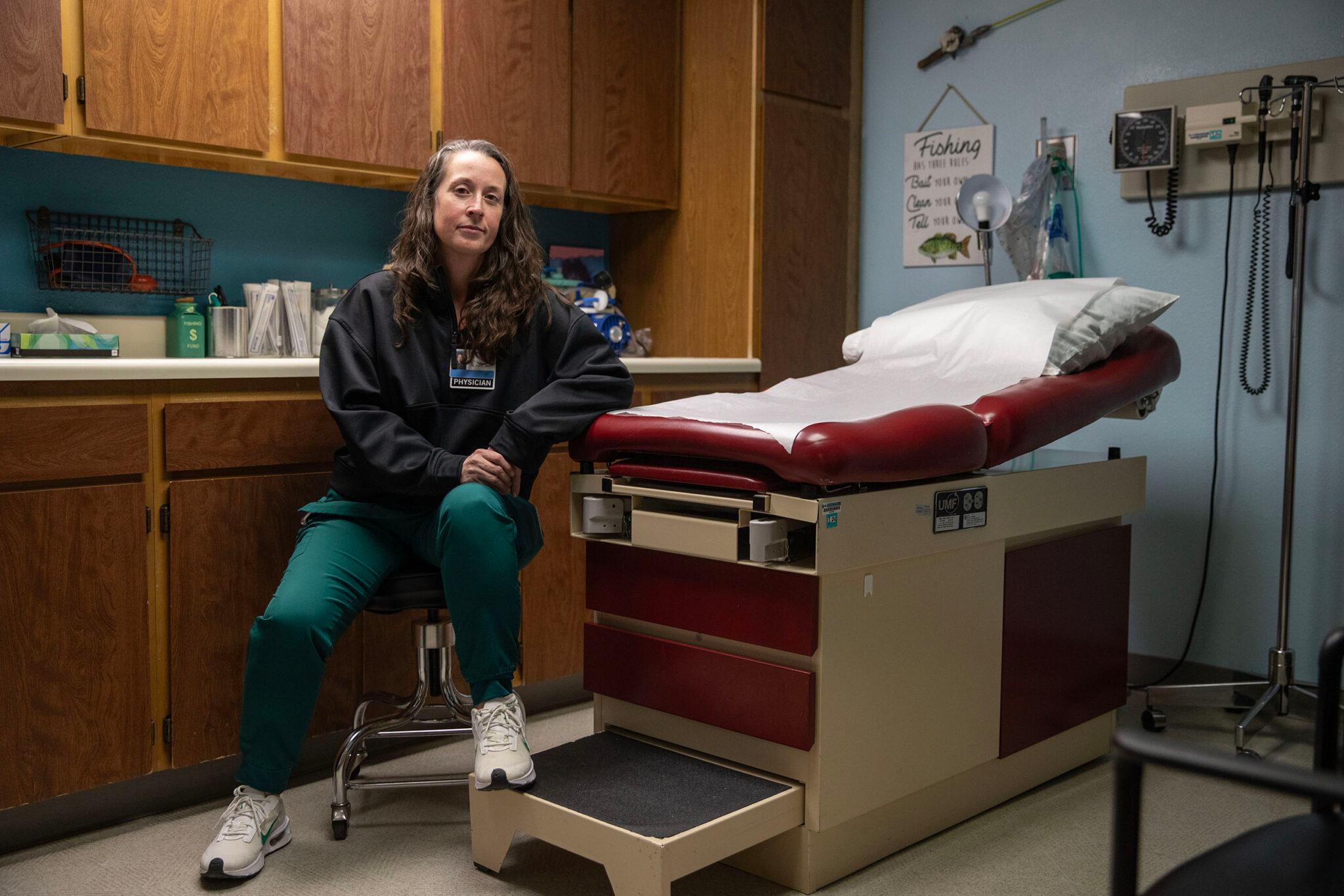 A woman in green medical scrub pants and a black sweater sits in an exam room, leaning on an exam table of cream metal and red leather. Doctors tools — and a fishing rod — hang on the wall behind her.