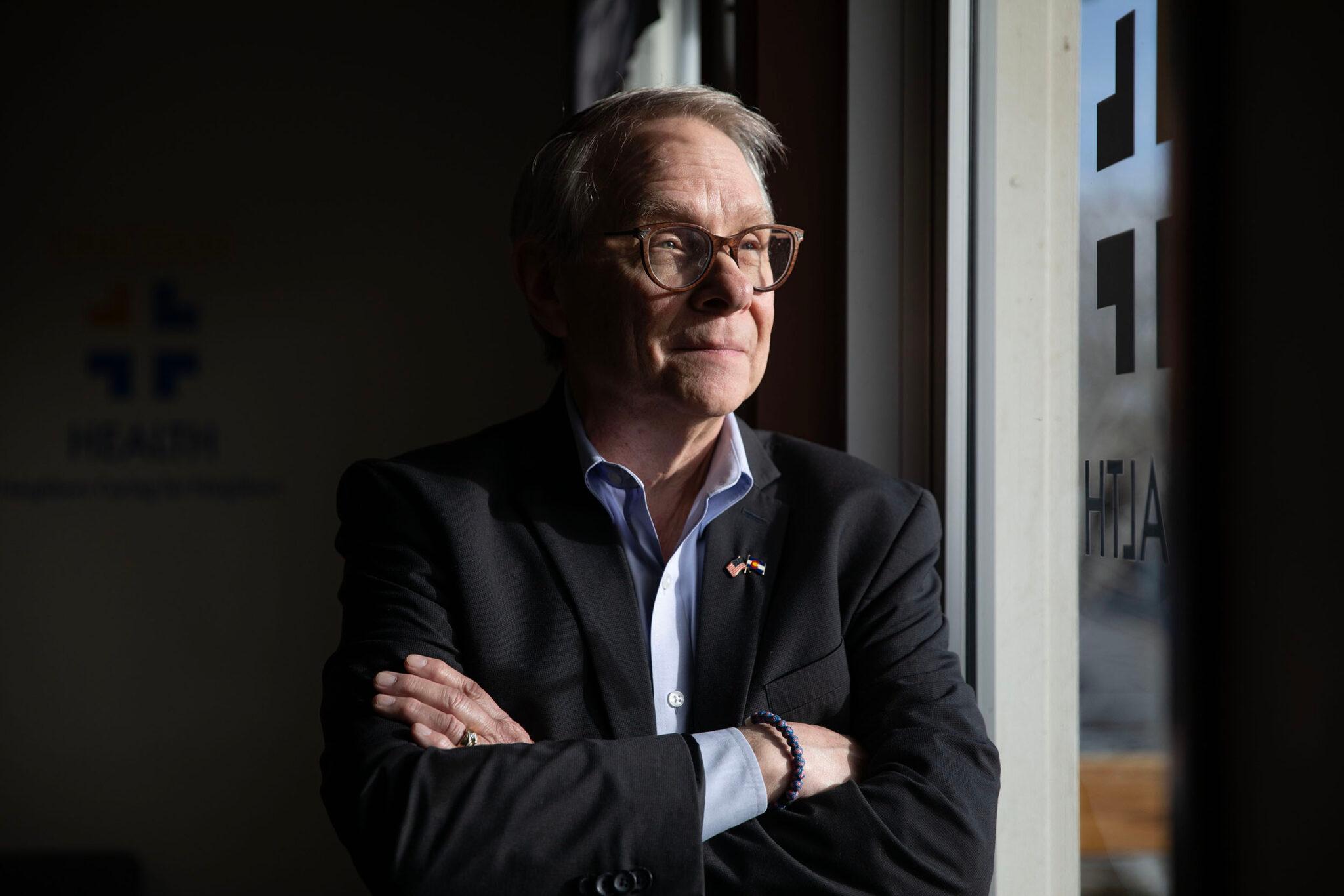 A man in a suit and brown-rimmed spectacles stands by a window and is lit dramatically by the incoming sunlight. He wears a slight smile on his face.