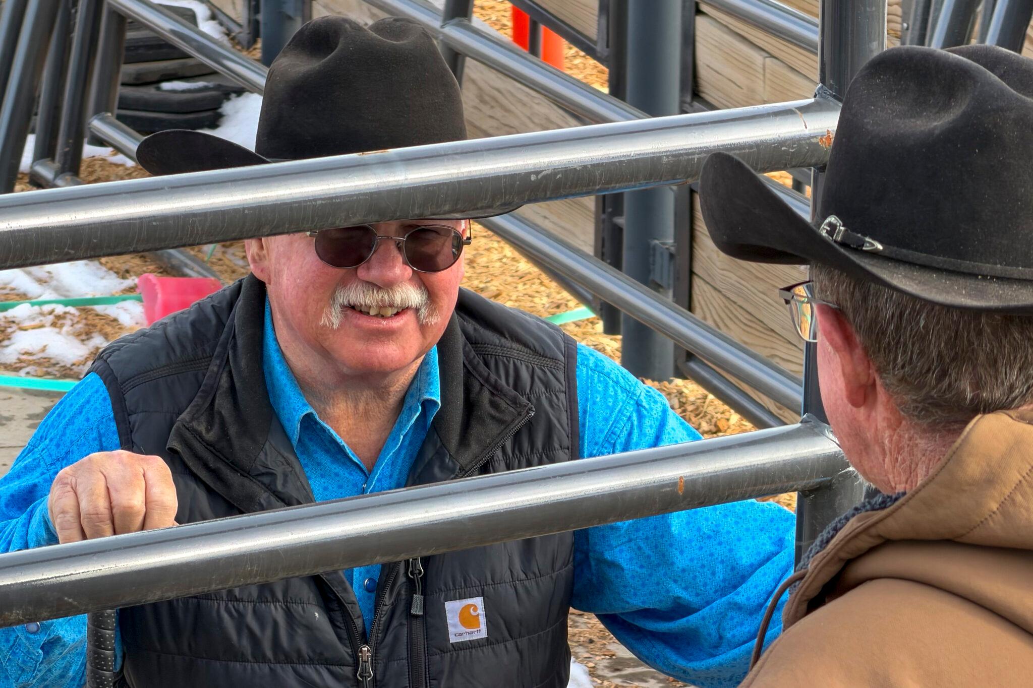 Two older guys in cowboy hats lean on a metal cattle fence as they chat.