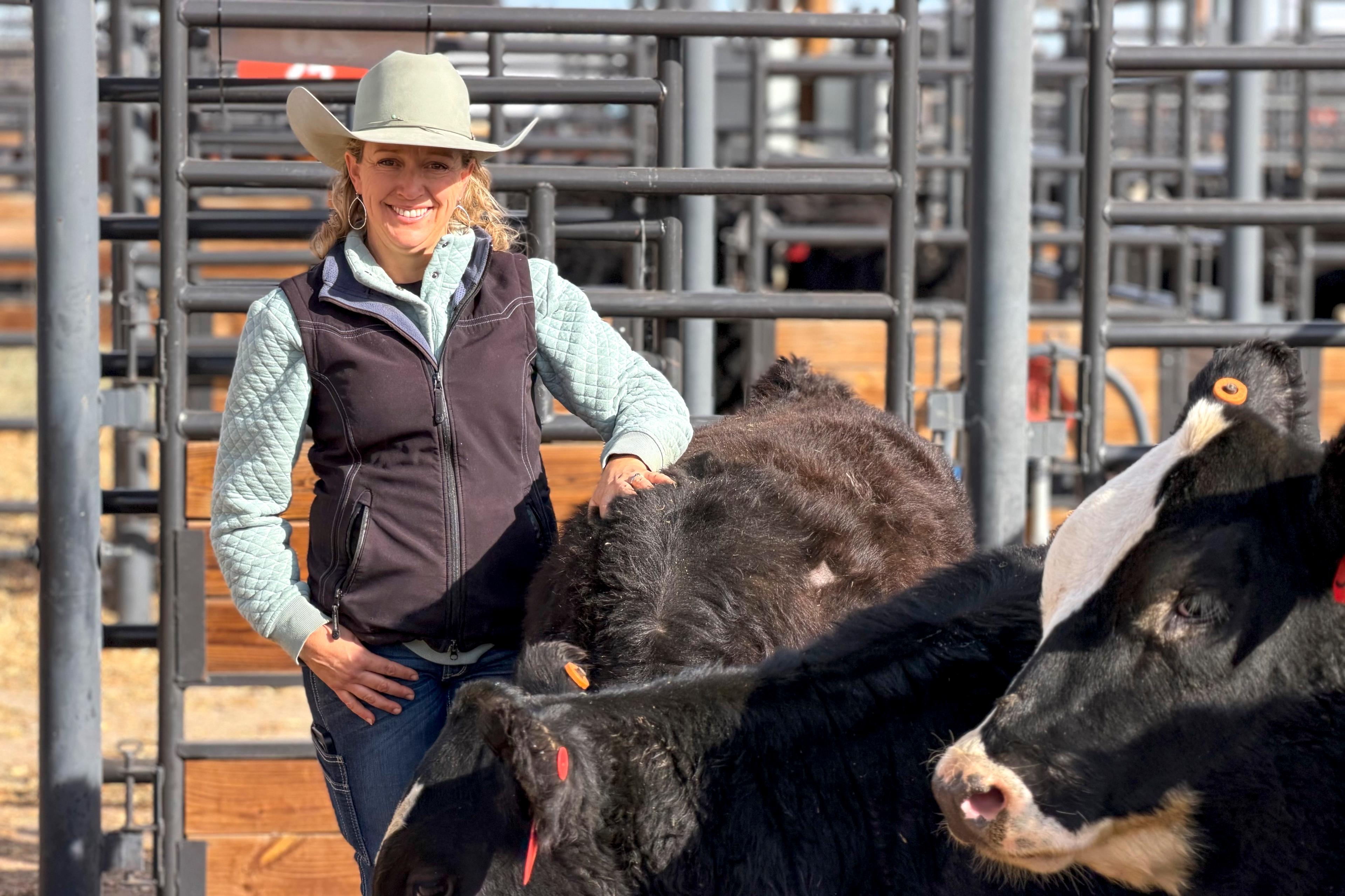 A woman in a vest and cowboy hat smiles at the camera as she pets one of a few cows gathered by her in the foreground.