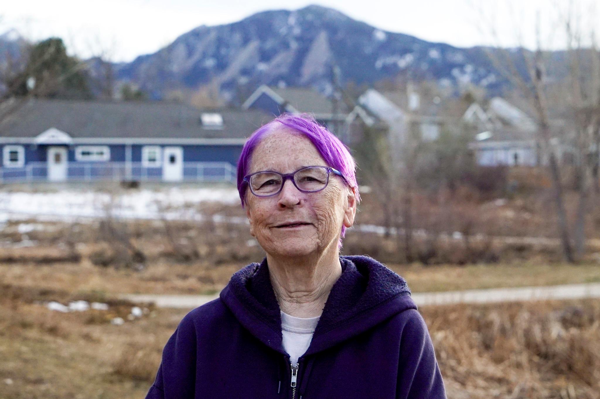 A woman with purple hair and wearing a purple hoodie looks into the lens, with blue buildings and blue mountains behind her.