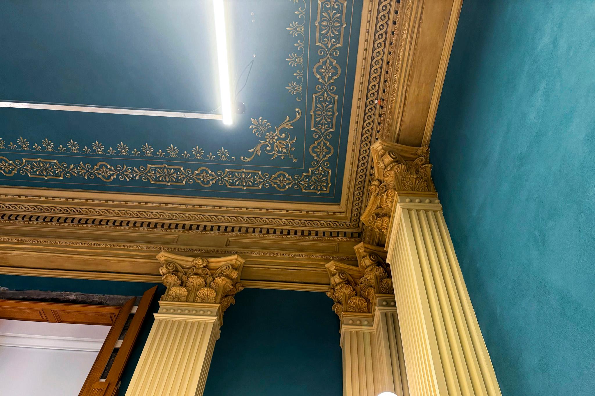 A look up at a blueish-green ceiling gilded in gold trim and held up by ornate columns.