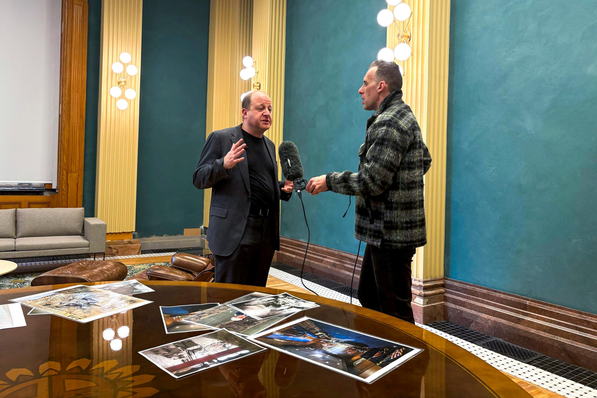 A man in a blue suit jacket speaks to a man holding a fuzzy microphone, in a room of teal walls lined with ornate gold columns.