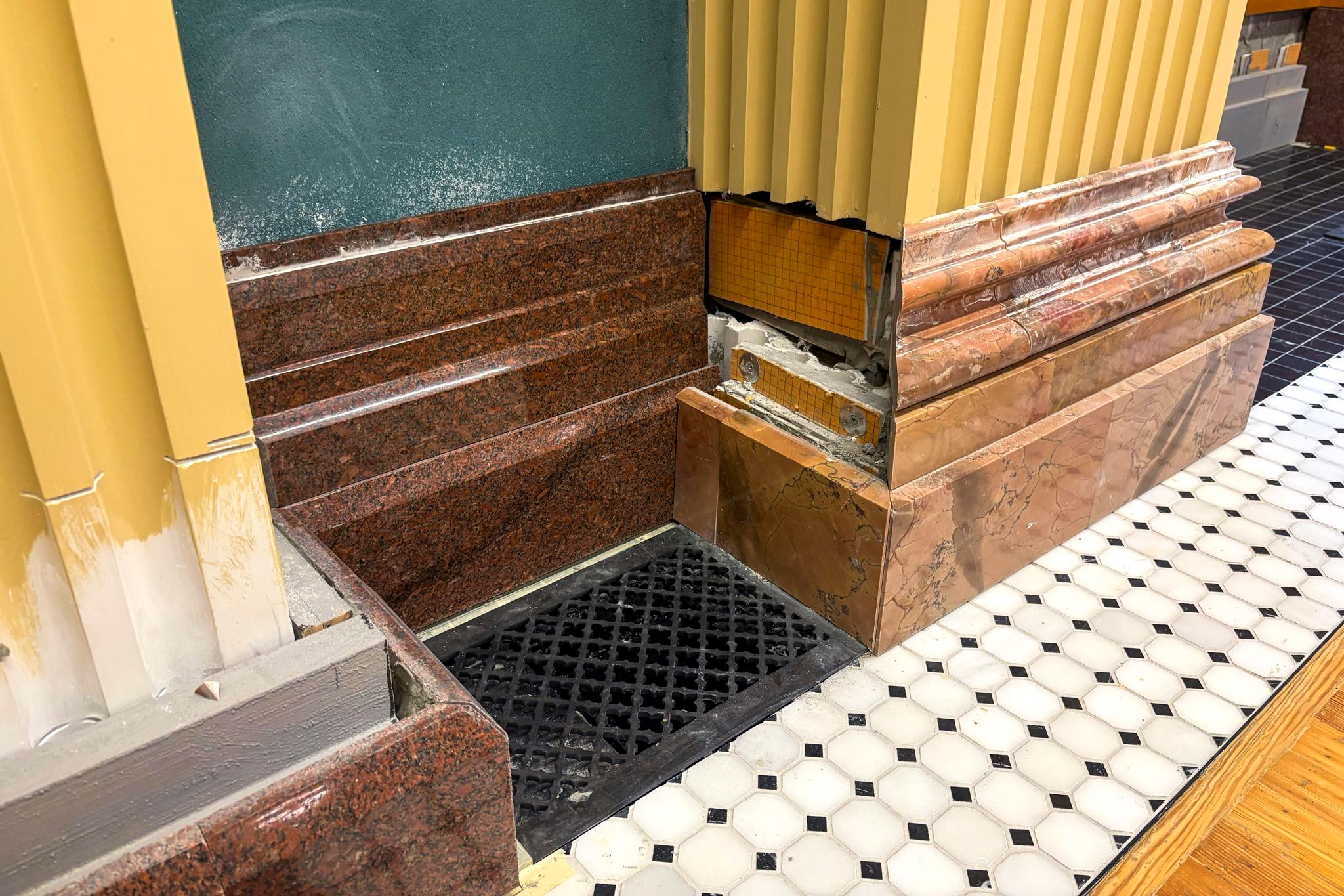A grate sits in the floor between two gold columns. A piece of the red rose quartz stone baseboard is missing above it, looking rather shoddy.