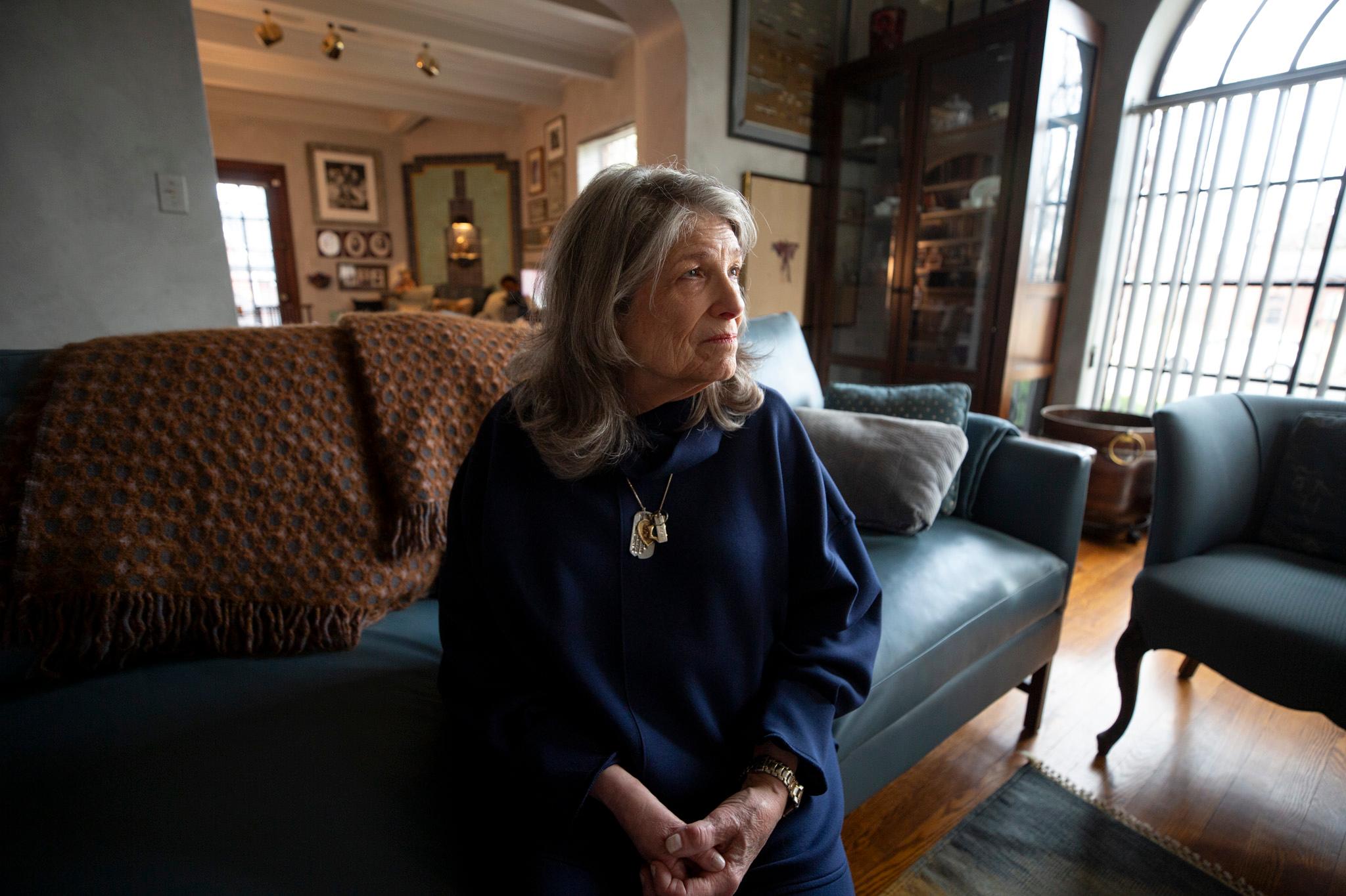 A woman in a navy cloak sits on a green couch by a bright, tall window. She looks out of frame, as if in deep thought.