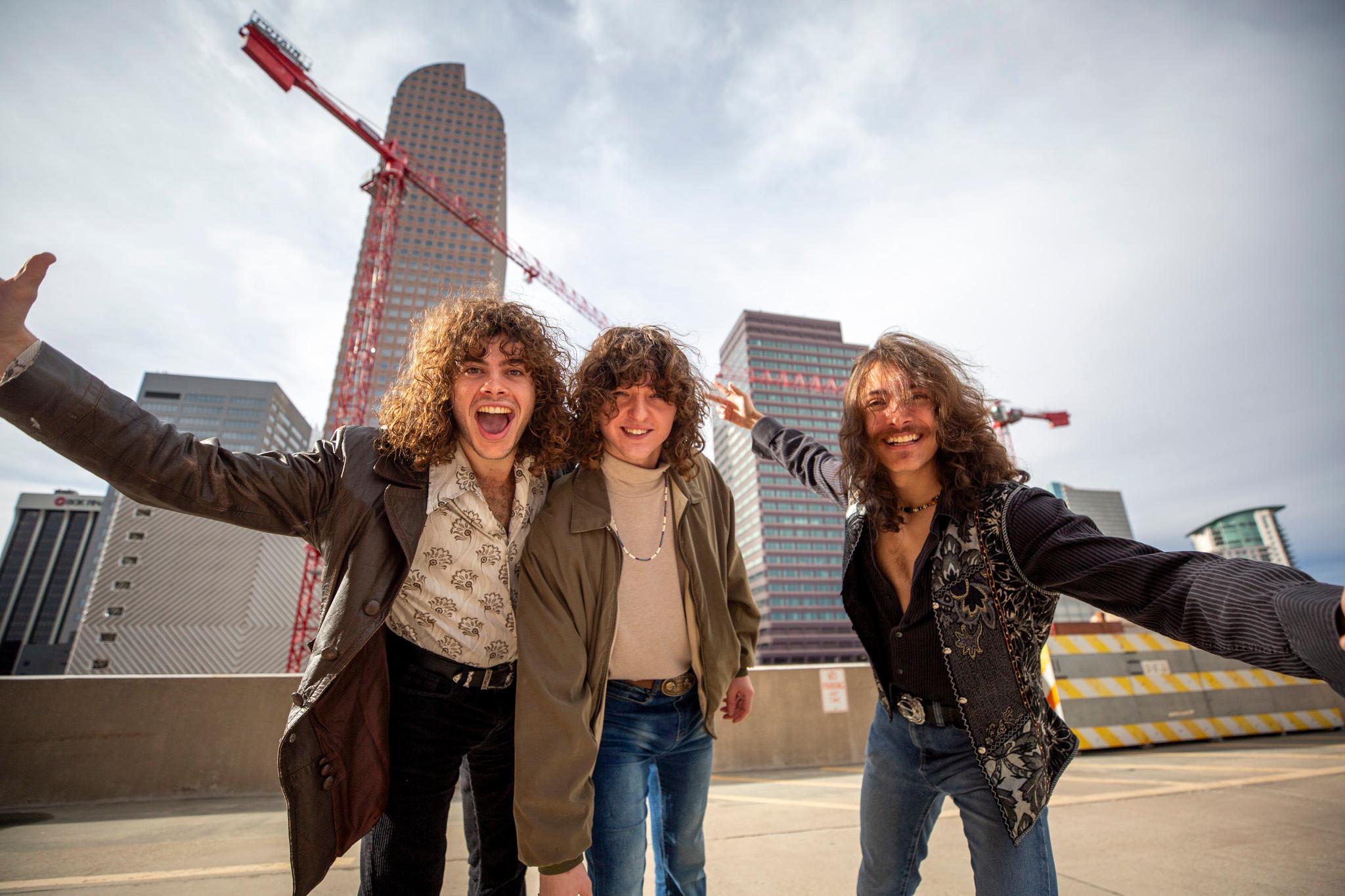 Three dudes with flowing hair and clad in leather jackets lean down into the lens, two with arms spread out wide, as skyscrapers rise behind them.