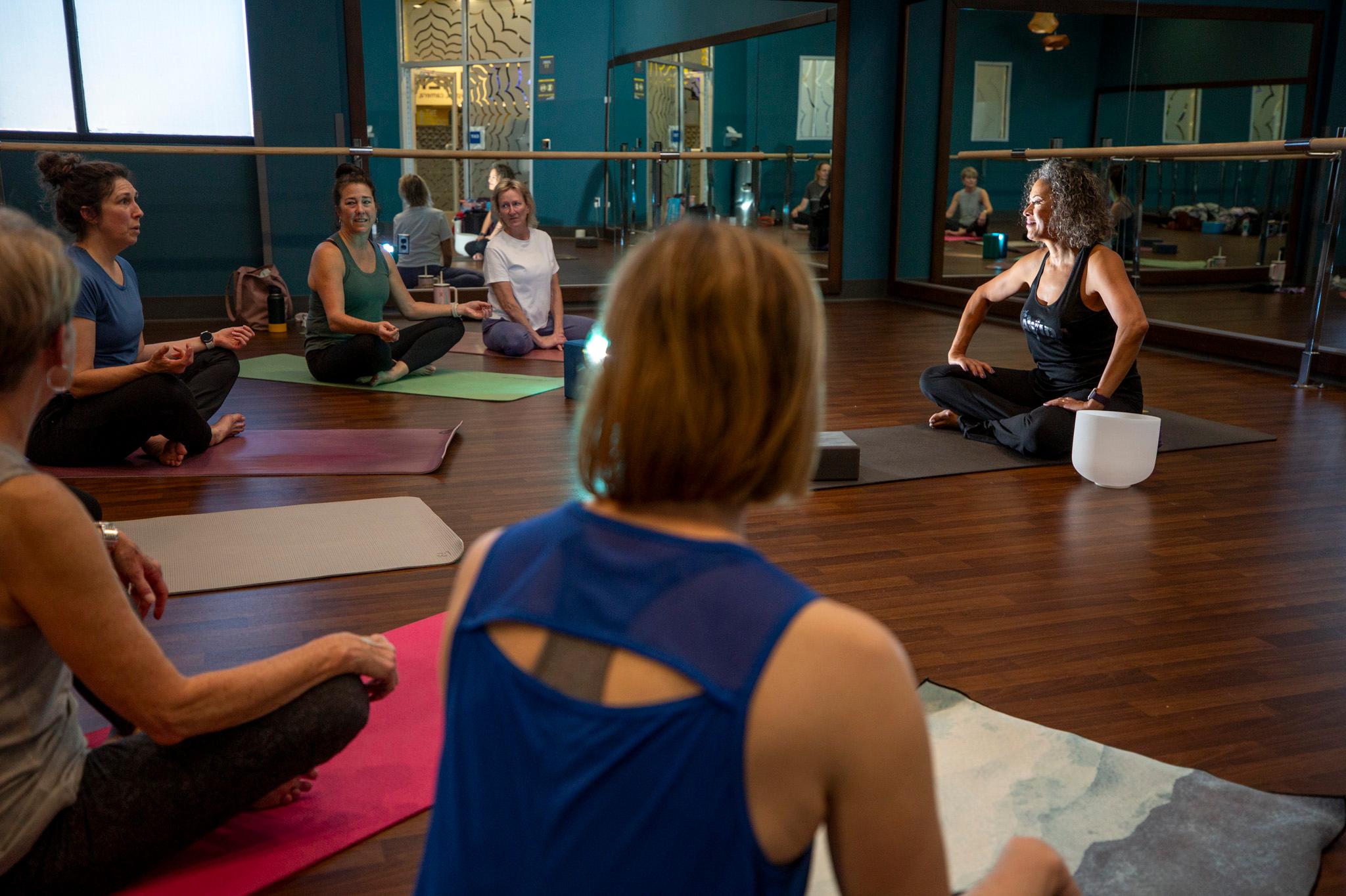 image shows women sitting on colorful mats on a wood floor surrounded by mirrors in a yoga pose. The instructor is in black seated on the right side of the photo.