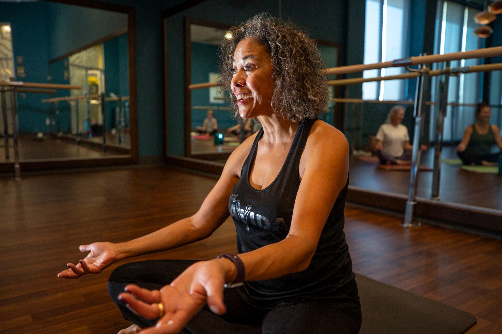 Photo shows a woman wearing a black tank top in a yoga pose with her hands outstretched. She is in front of walls of mirrors in a yoga studio.