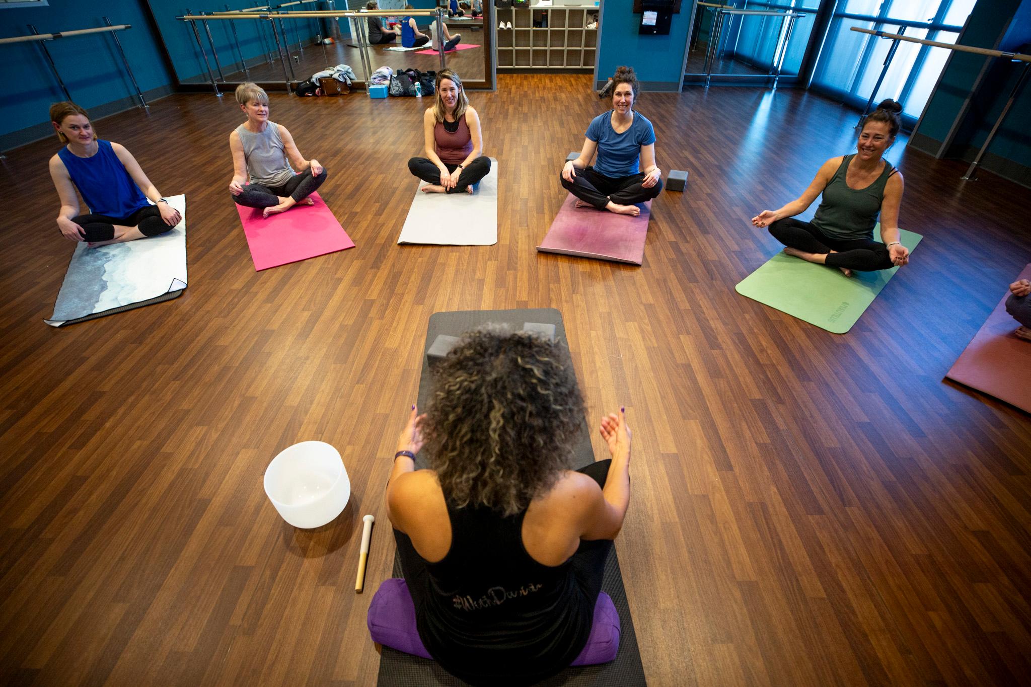 Photo shows a semi-circle of women in yoga poses at the top of the image and and the bottom of the image is the instructor, also in a yoga pose, facing them.