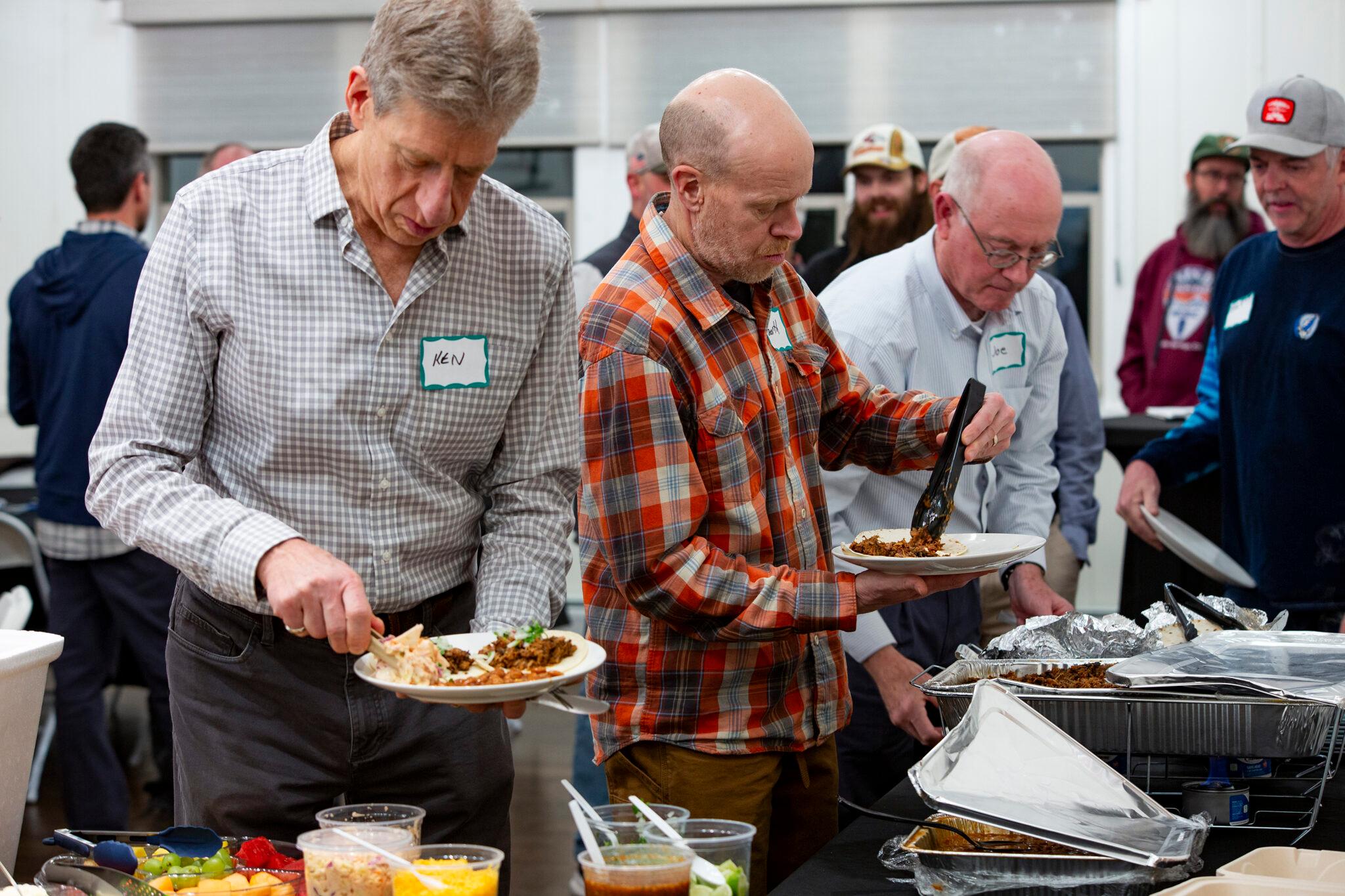 Three older men wearing nametags stand in line above a buffet of food, each shoveling tacos and meat onto their plates.