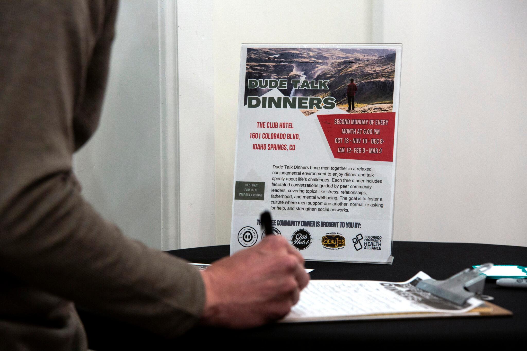 A sign on a table reads "dude talk dinners." A hand reaches in from the left side of the frame, signing a clipboard on the table below the sign.