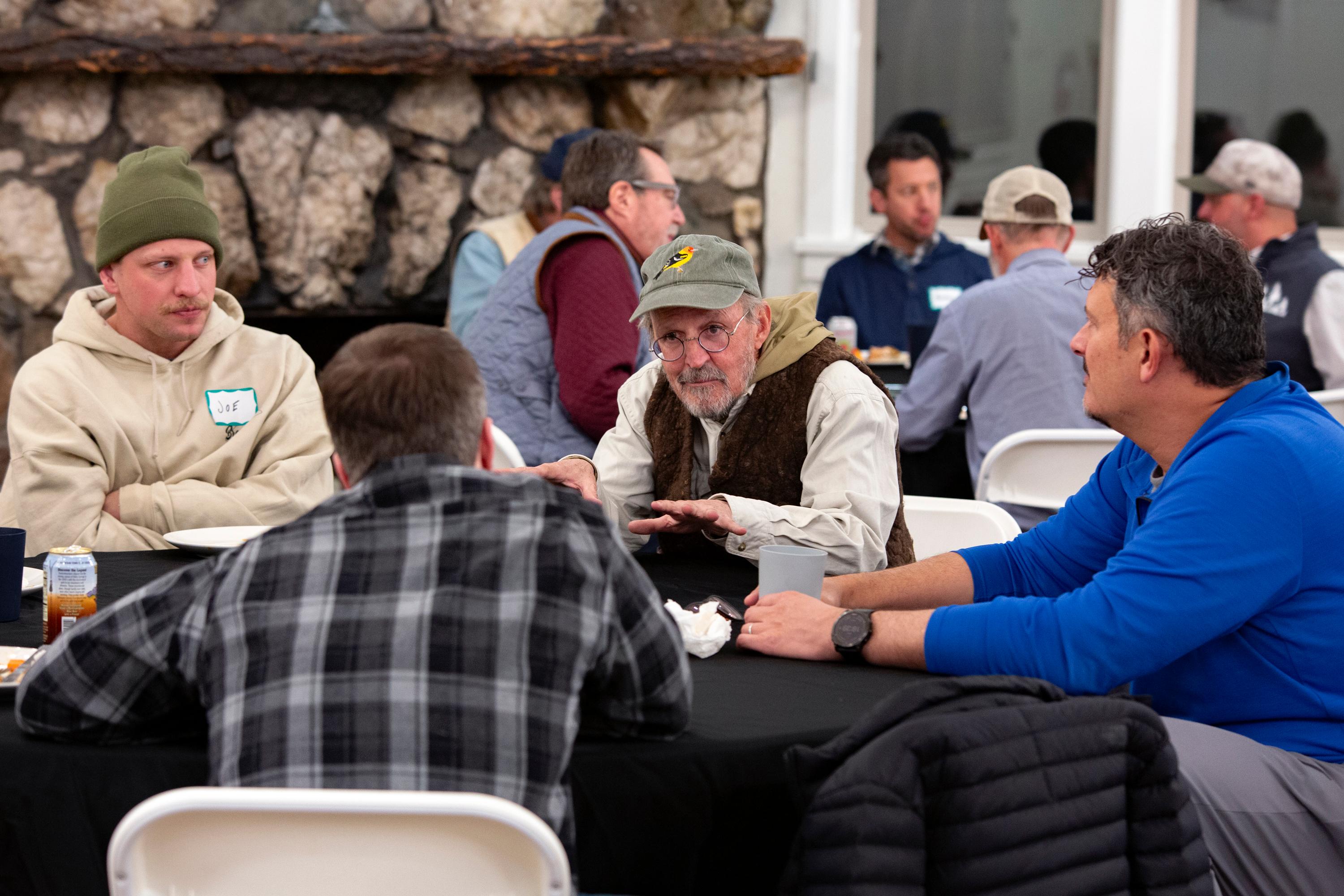 Two groups of guys sit on plastic chairs arranged around linen-covered tables, looking intently at each other in clearly deep conversation.