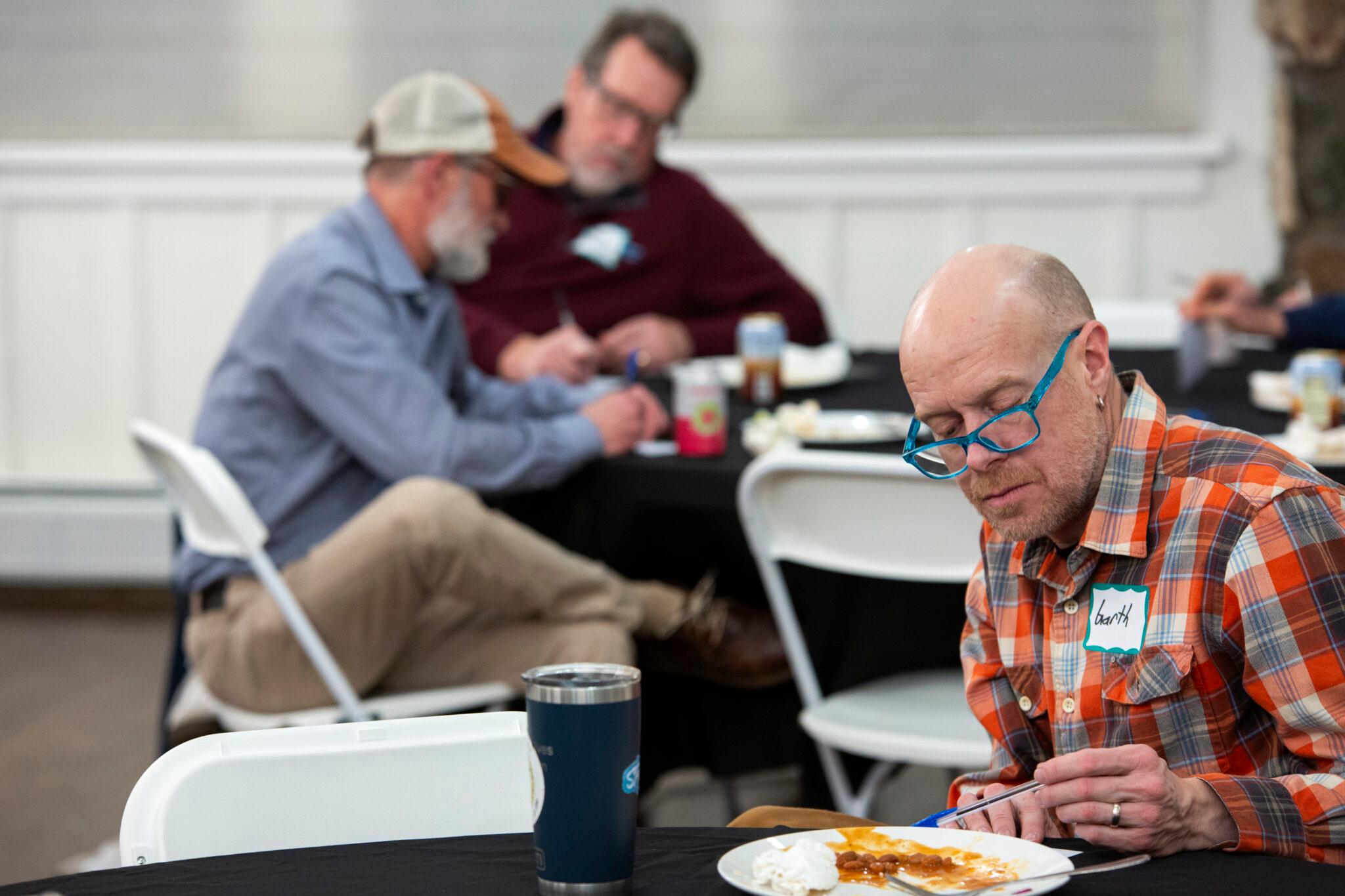 A man in bright blue spectacles points a pen at a piece of paper on a table before him, concentrating on an apparent assignment. Others, out of focus behind him, do the same.