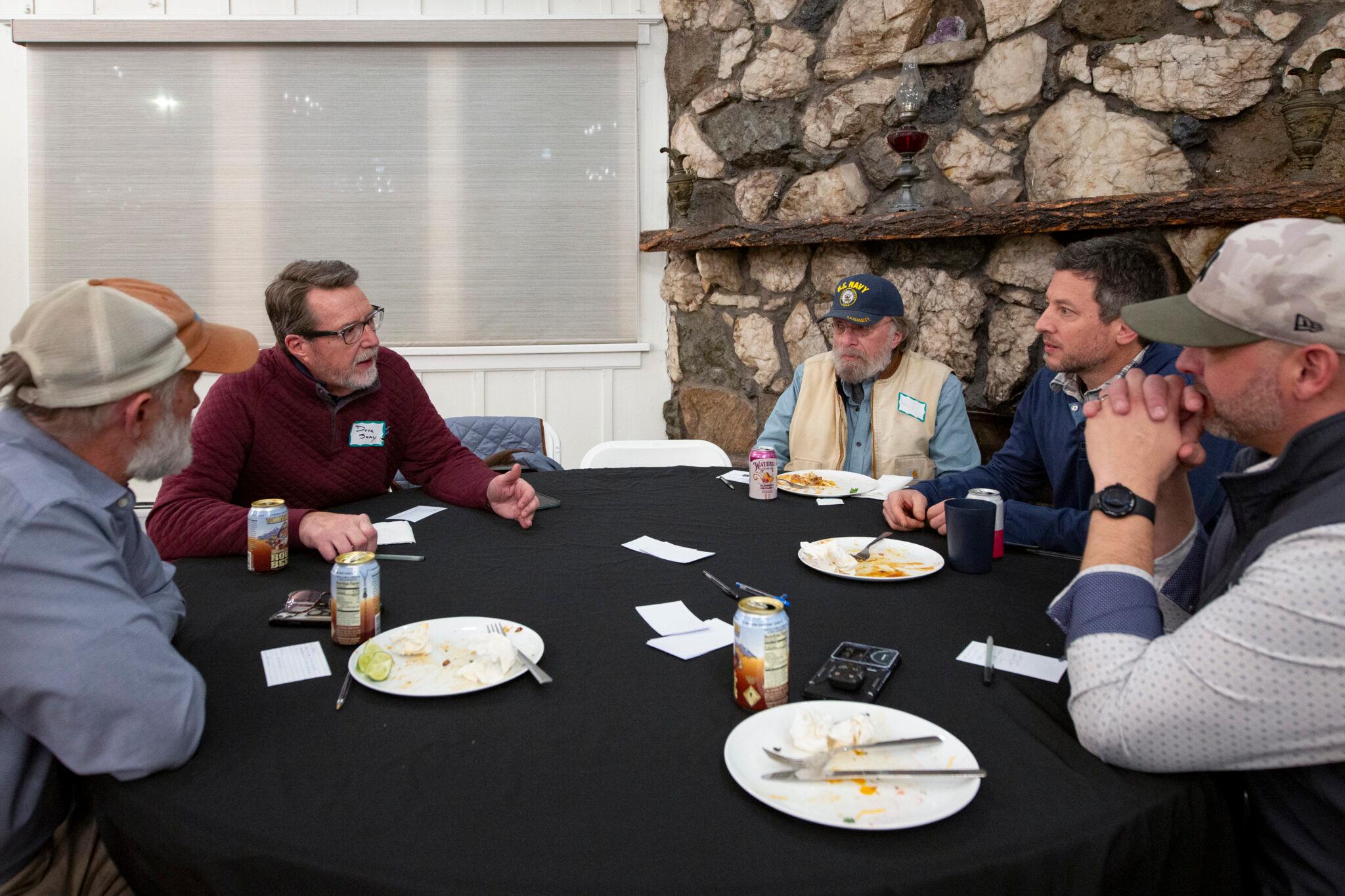 A group of men sit in plastic chairs around a linen-draped table, concentrating on their conversation. One man in a red shirt to the left speaks — the camera angle makes it look like we're seated with him.