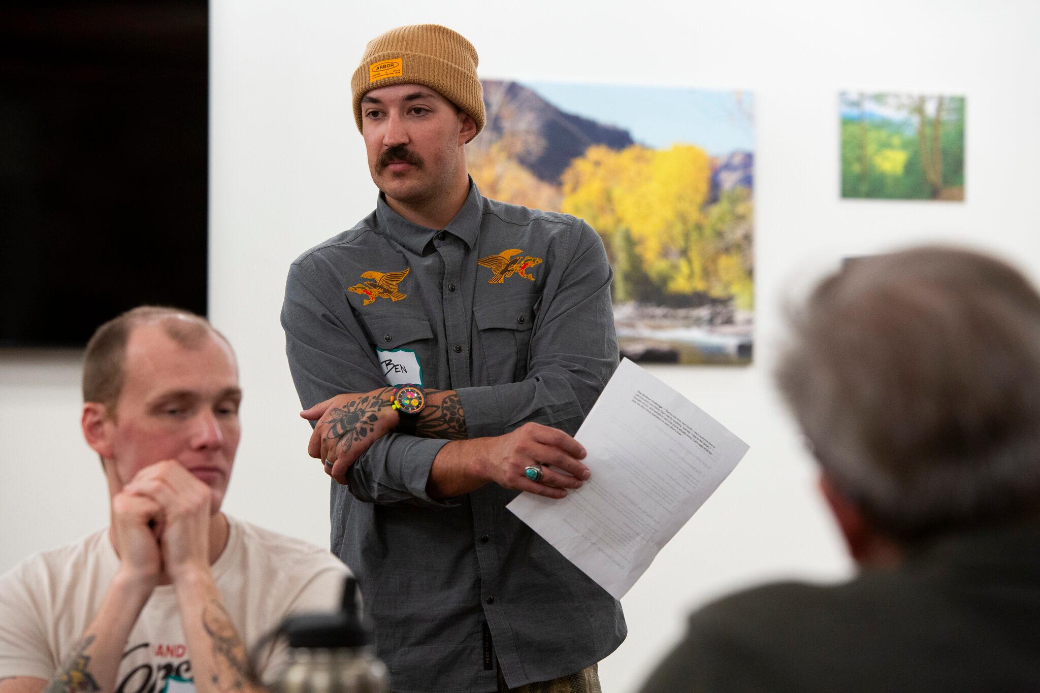 A mustached man in a knit beanie and buttoned work shirt stares into a room, past the camera. In the foreground are two men, out of focus, who appear to be sitting and talking.