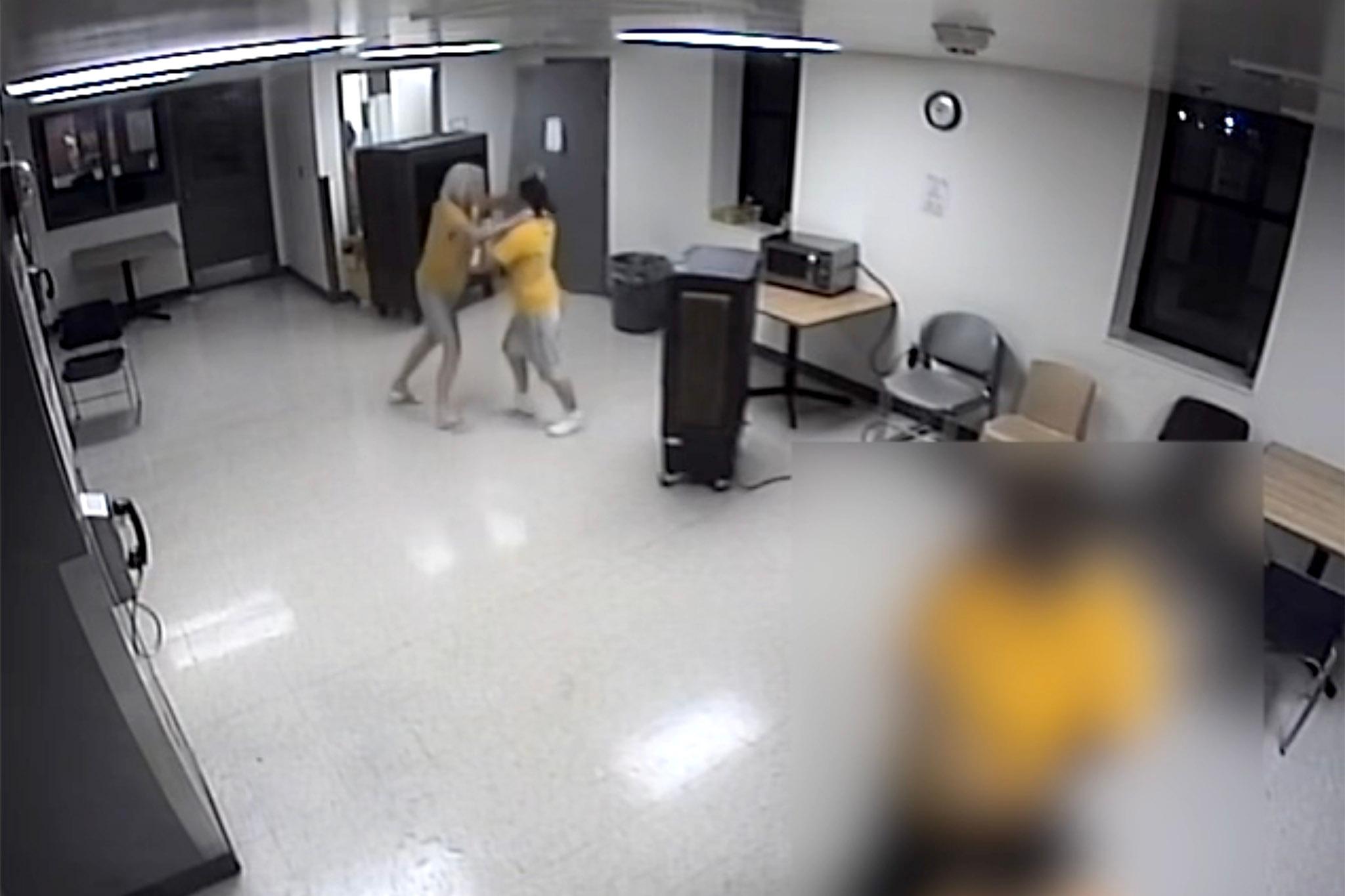 A grainy, wide-angle image that clearly comes from a security camera, depicting two women in yellow in a white room. One appears to have her hands around the other's neck. Someone in the foreground has been blurred out.