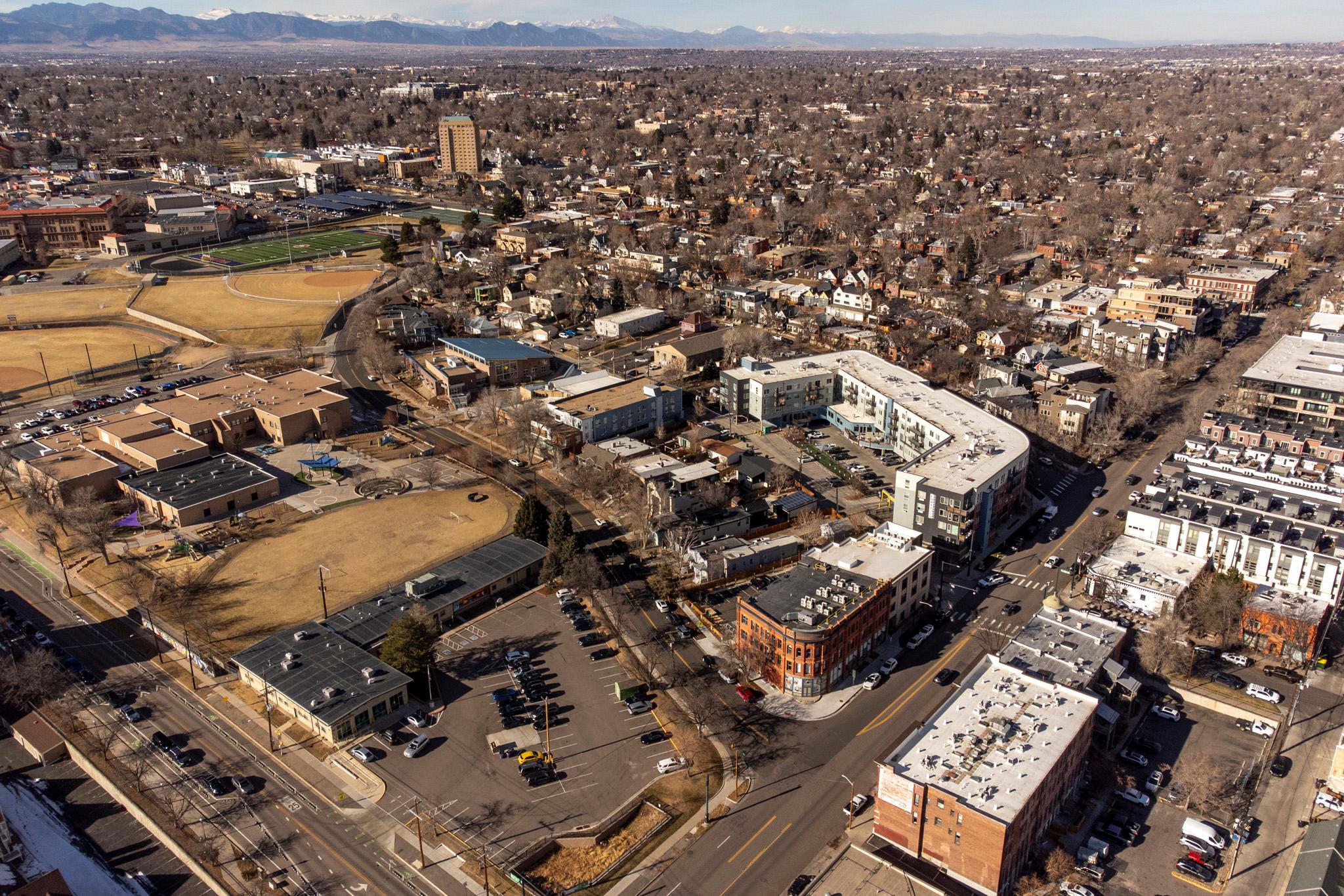 An aerial view of a red brick building on the corner of an intersection with a sprawling neighborhood and sports fields behind it. Mountains rise on the horizon.