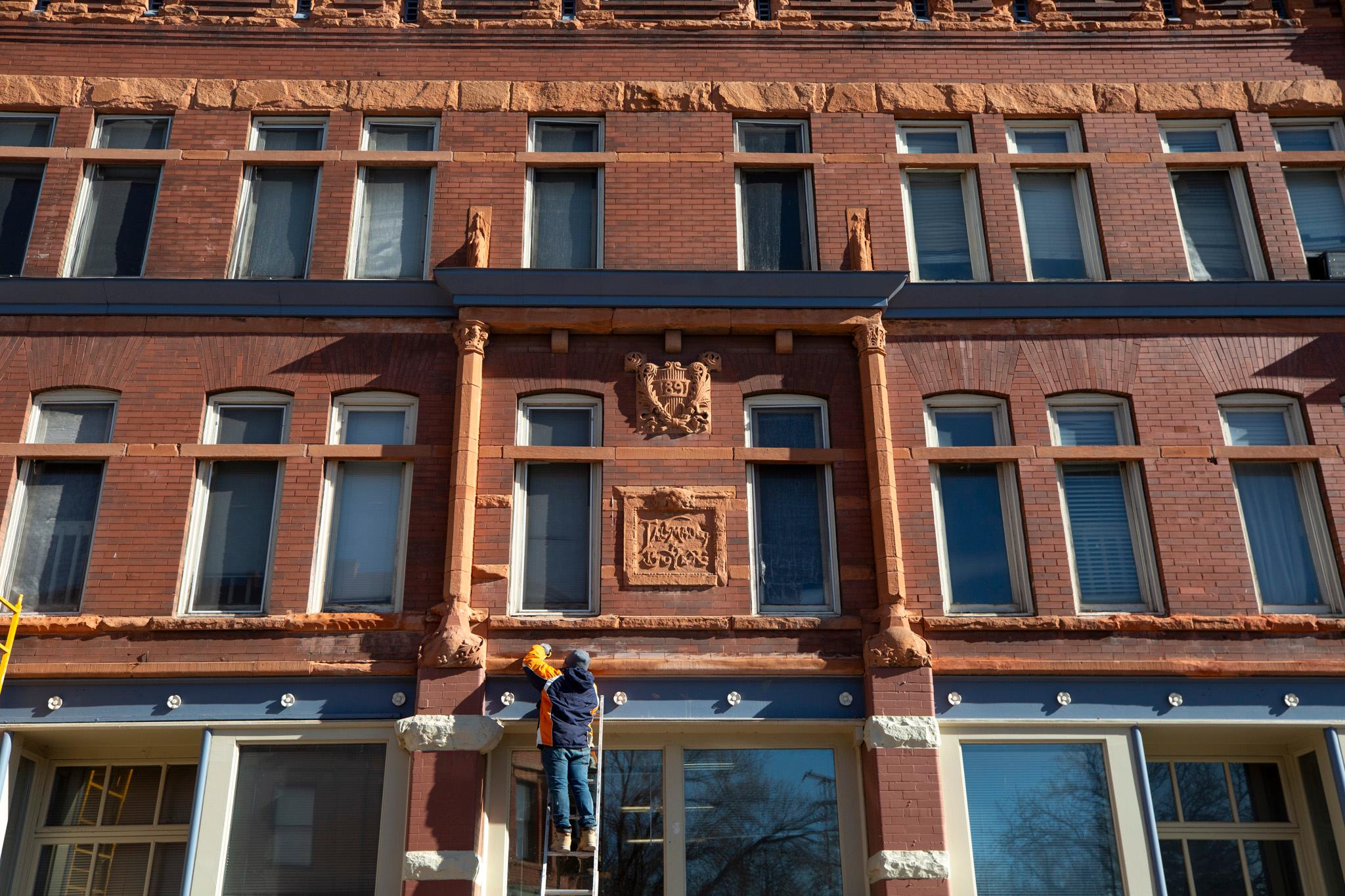 A man stands on a ladder leaned up on a large brick building. A crest in the center reads "1891."