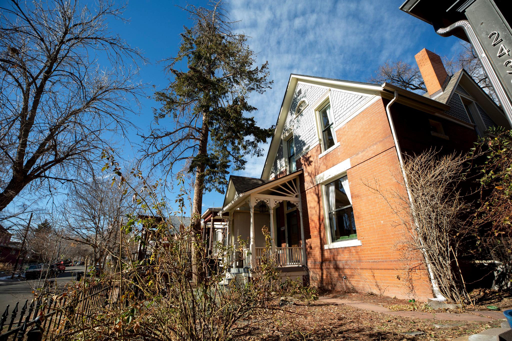 An orange brick building under a blue sky, with all kinds of weeds and bushes and trees in the yard around it.