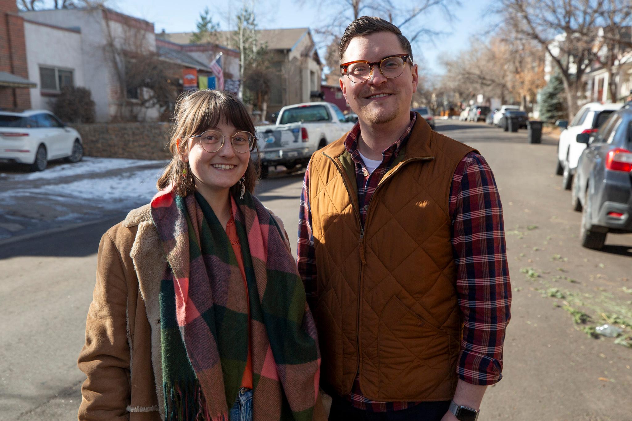A woman with a plaid scarf and a man in a plaid shirt and vest stand in a street and smile at the camera.