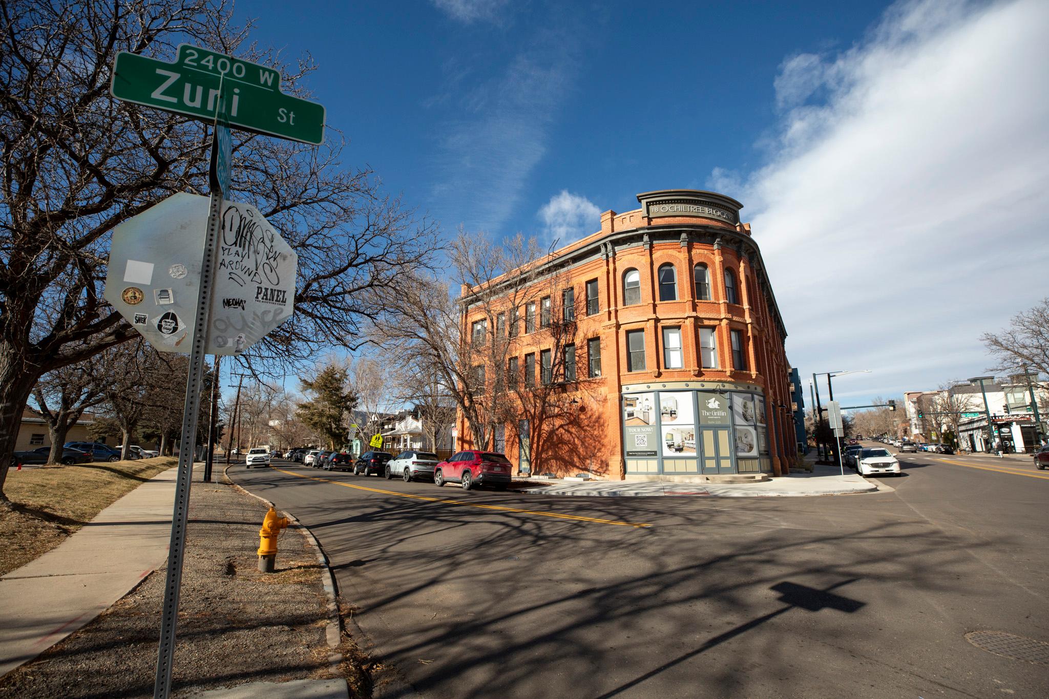 A red brick building sits on a corner under a blue sky. A sign in the foreground reads "Zuni St."