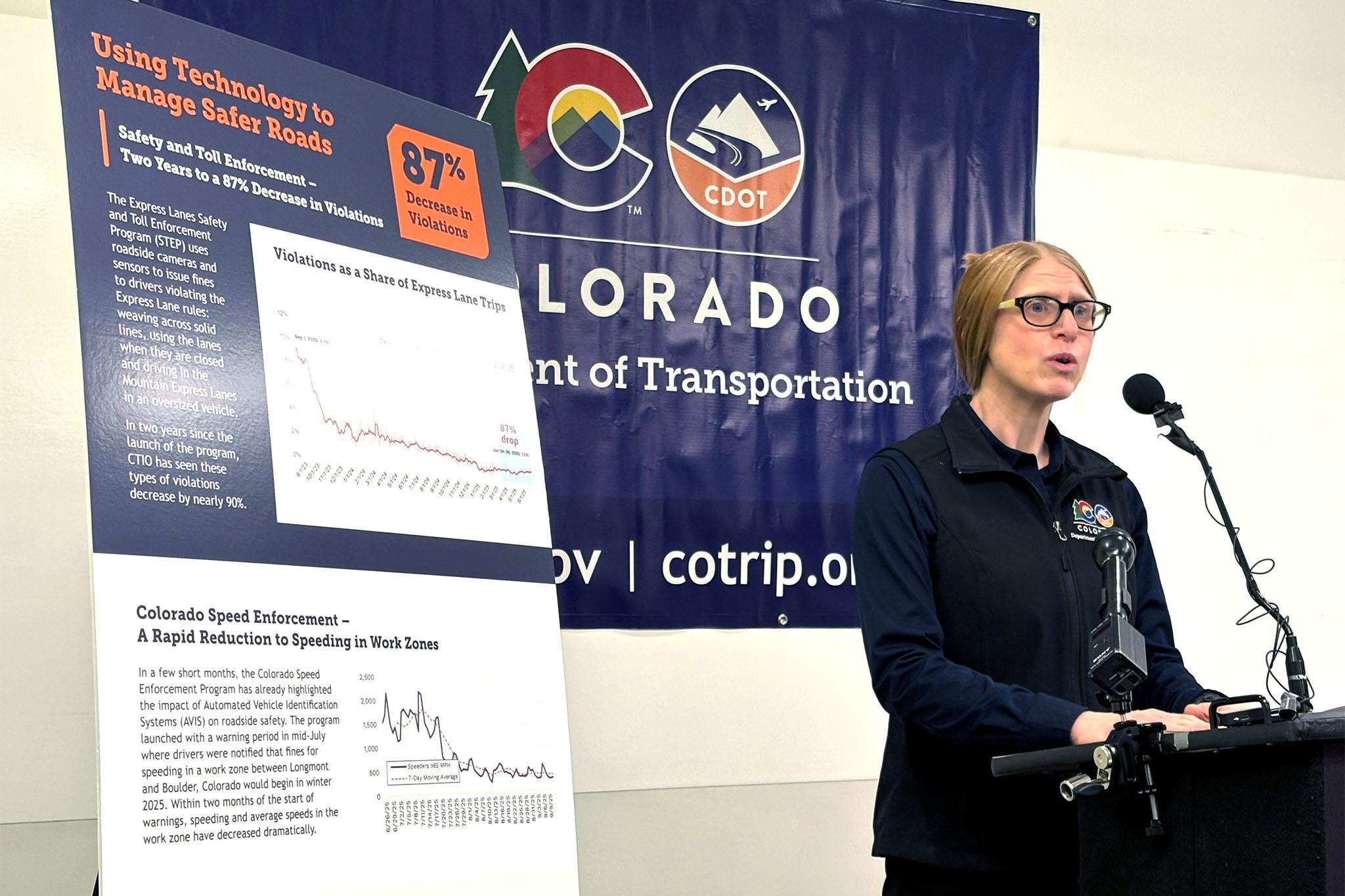 A woman in a black vest and glasses stands next to line charts that show express lane violations and work-zone speeding violations that are generally on the decline. She speaks into microphones at a podium.