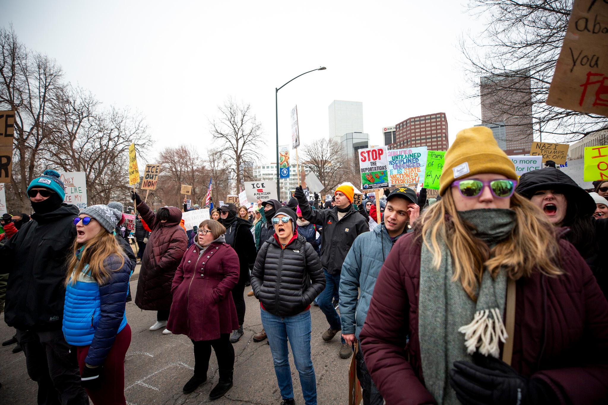 A large crowd stands in a street. Many are yelling, and more hold signs. One reads "STOP THE DEPORTATIONS."