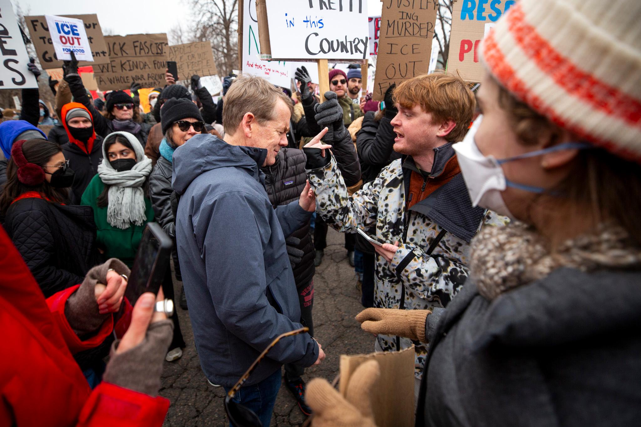 A man in a blue coat winces as a younger man points a middle finger right in his face. They're surrounded by a crowd of people, many of whom are holding signs.