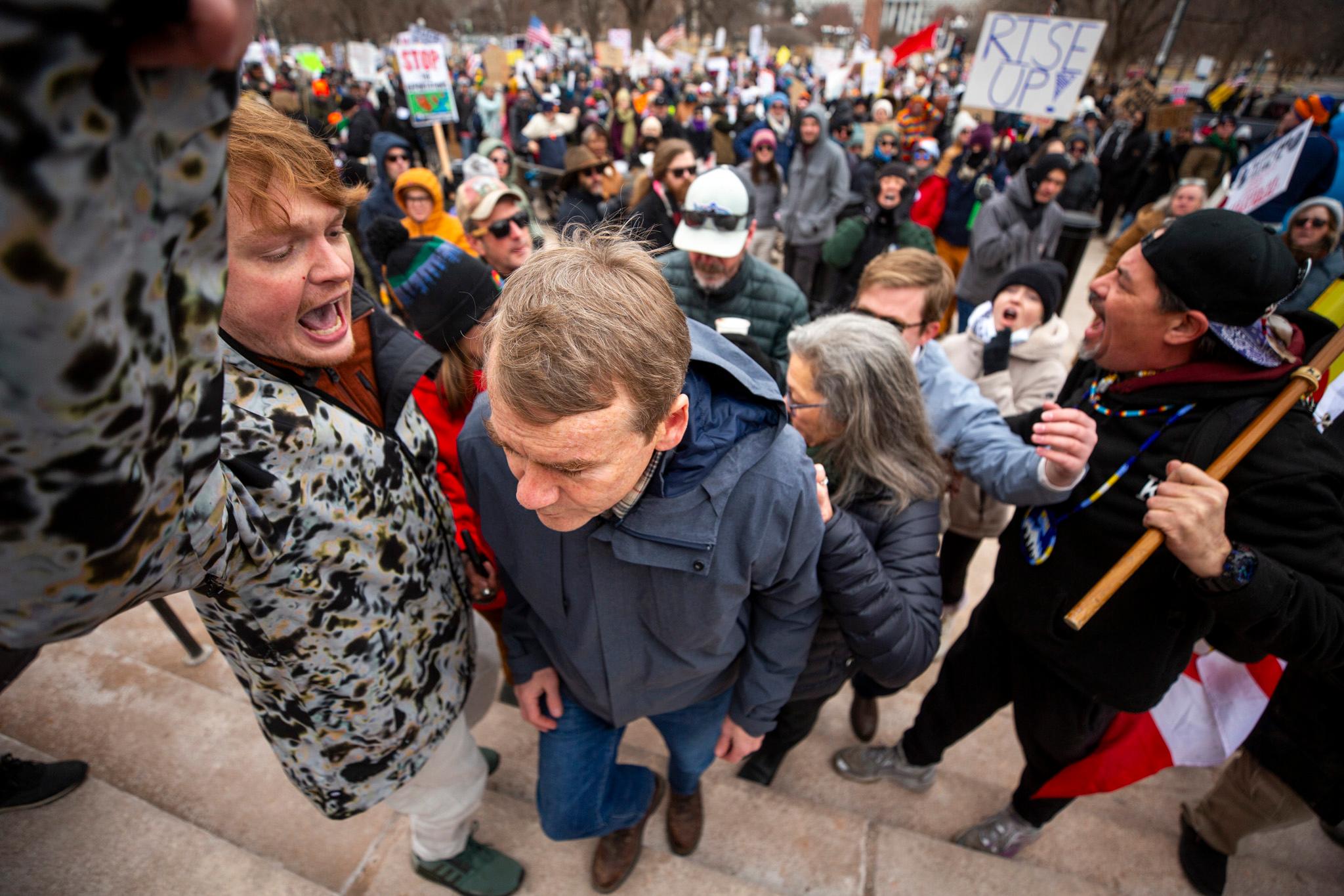 A man in a blue coat attempts to pass a younger man who is yelling right in his face. Behind them is a large crowd.