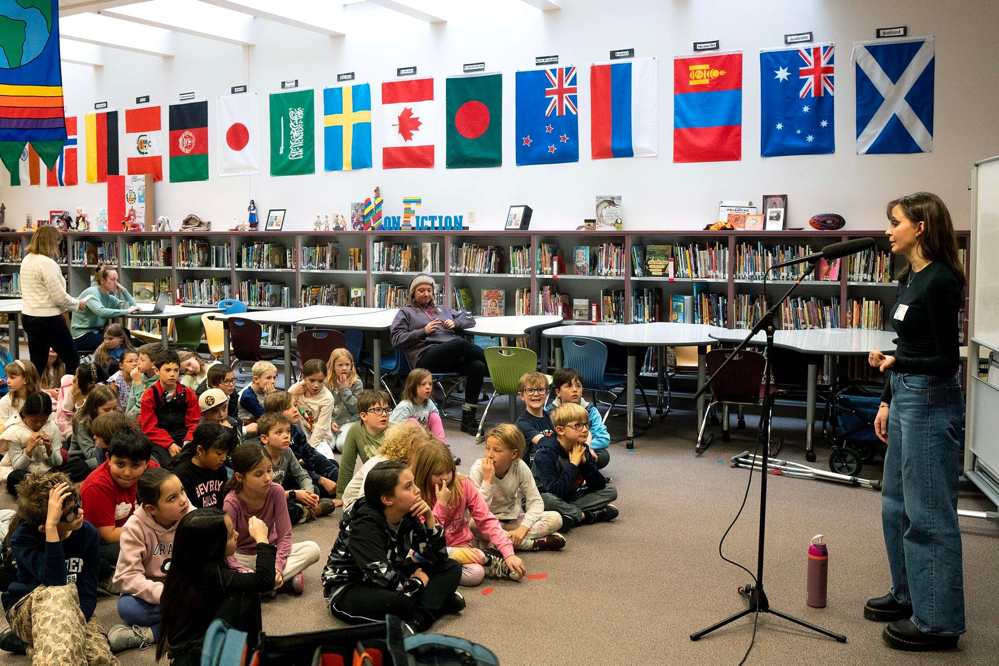 A kids sit in a crowd on the floor as a woman in black speaks to them from a microphone. Behind them are shelves full of books and a dozen international flags hanging on the wall.