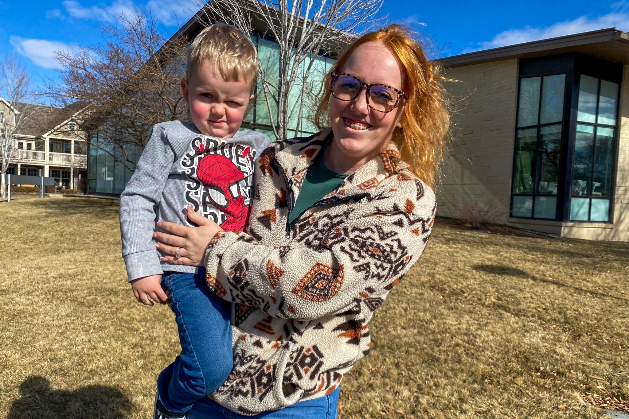 A woman in a patterned sweater smiles as she holds a little boy sporting a Spider-Man shirt. Behind them is a building of cream brick and blue skies.