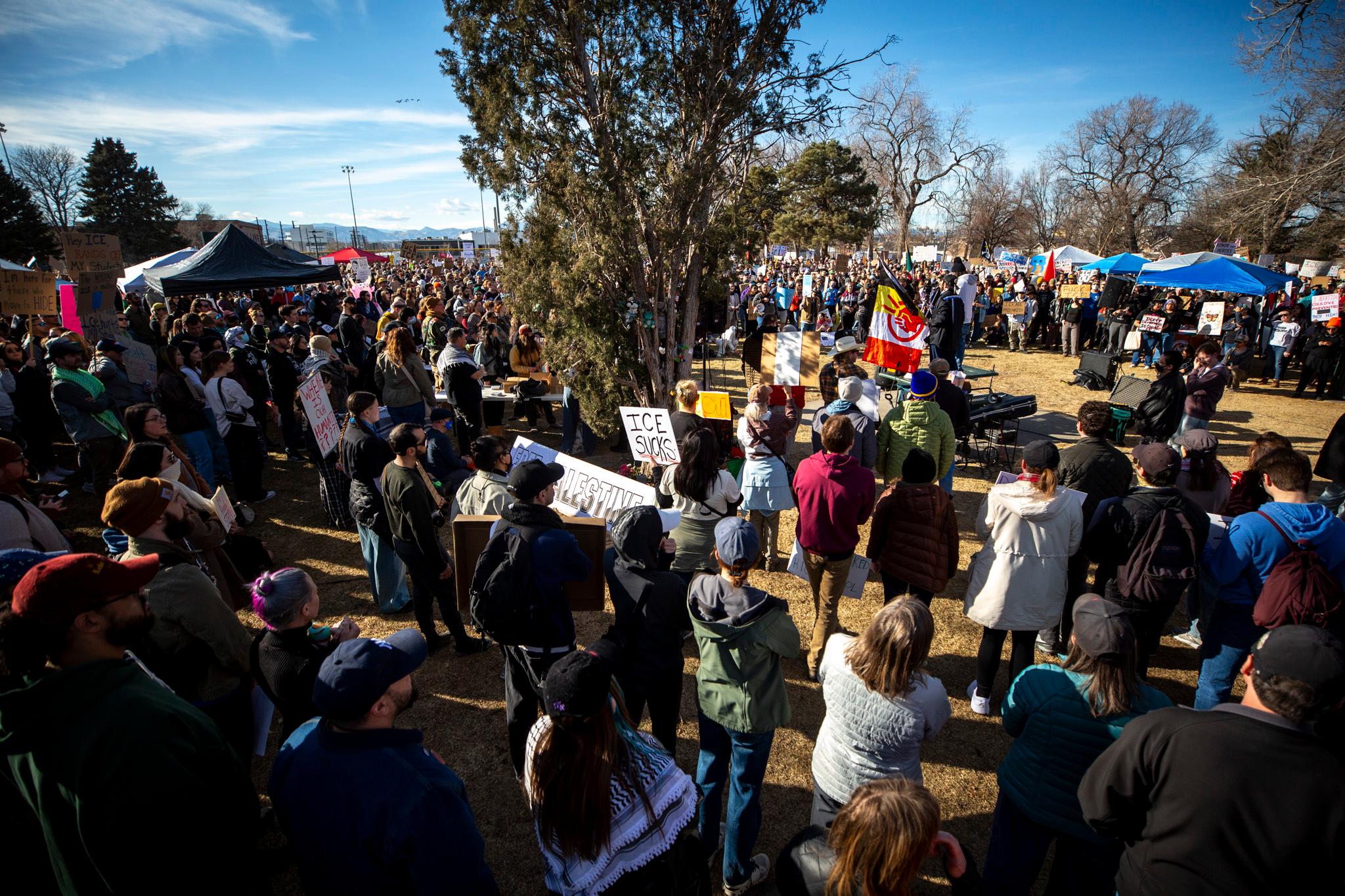 A large crowd stands under a bright sky. Many people hold signs.