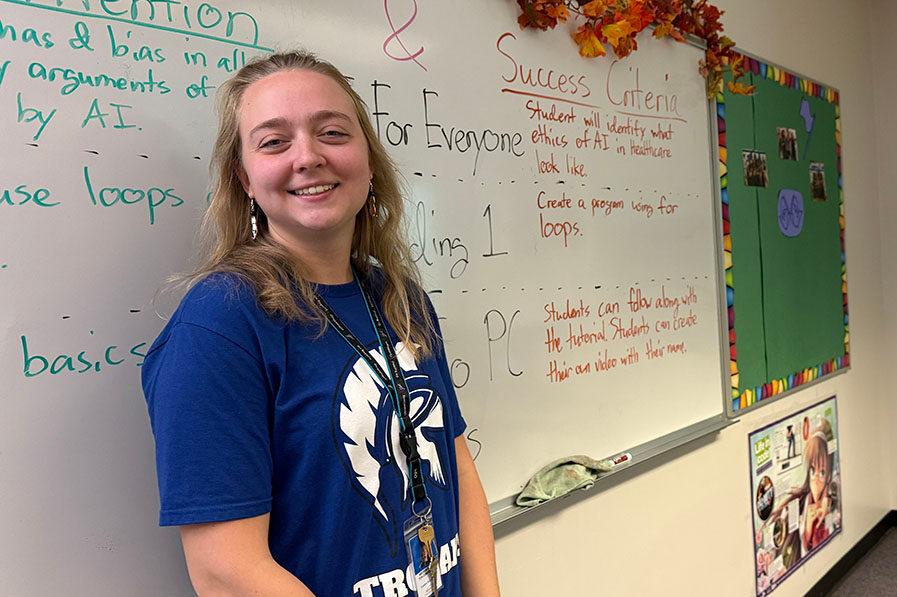 A woman wearing a blue shirt is standing in front of a whiteboard with writing on it. She is smiling for the camera.