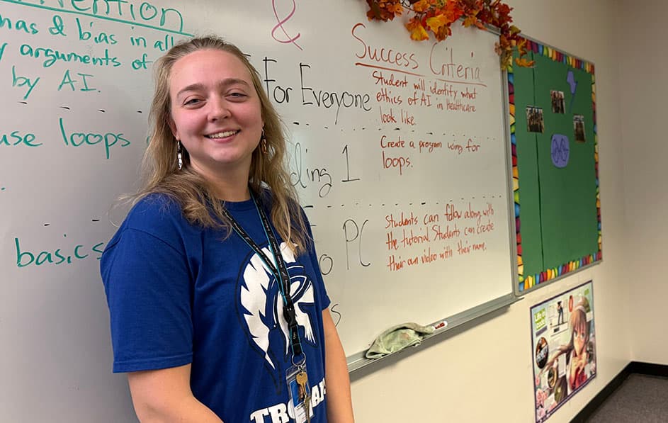 A woman wearing a blue shirt is standing in front of a whiteboard with writing on it. She is smiling for the camera.