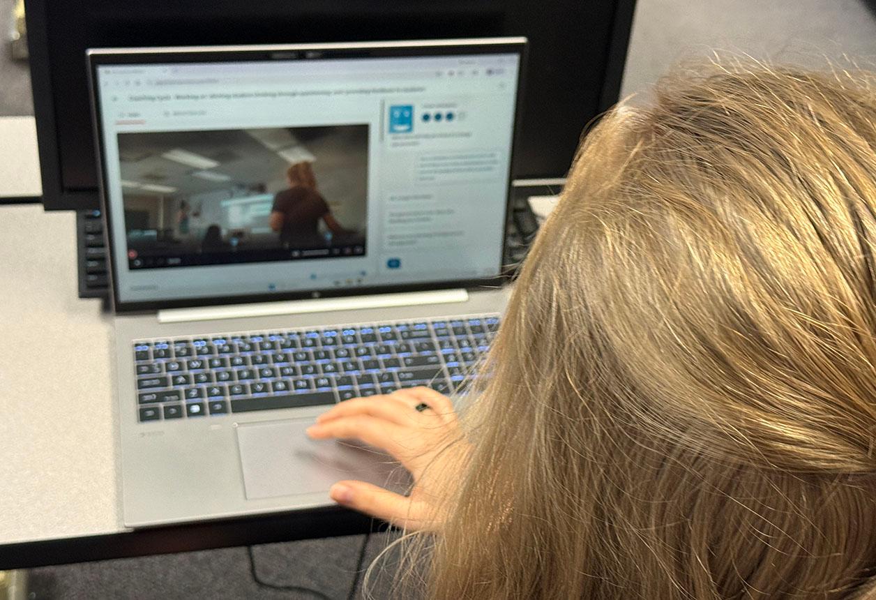 A person is using a laptop, which is displaying a video. The laptop is placed on a surface, and the person is focused on the content being displayed.