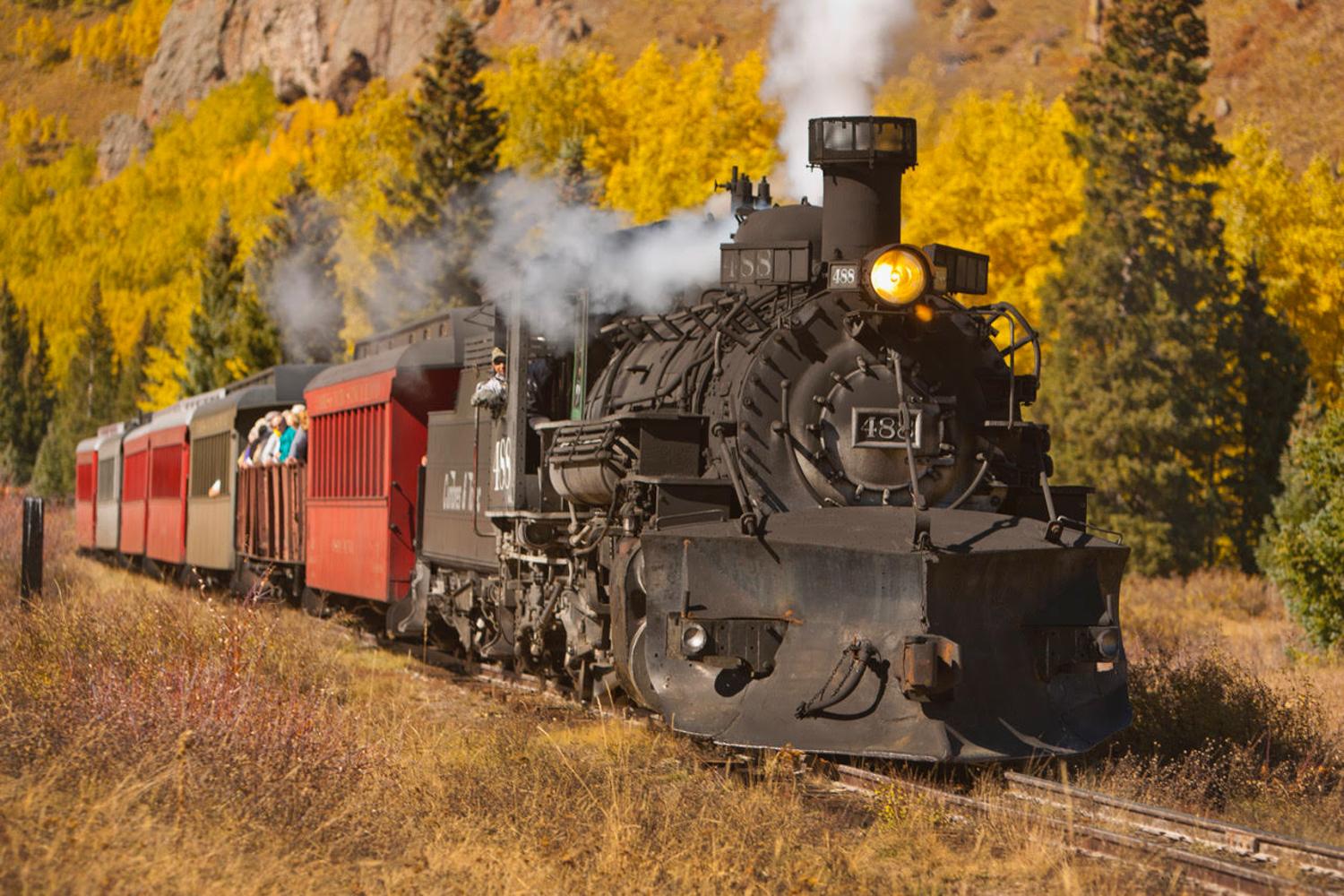 old-fashioned steam engine pulling several cars is traveling on tracks, with smoke billowing from its stack and there are people visible in the scene