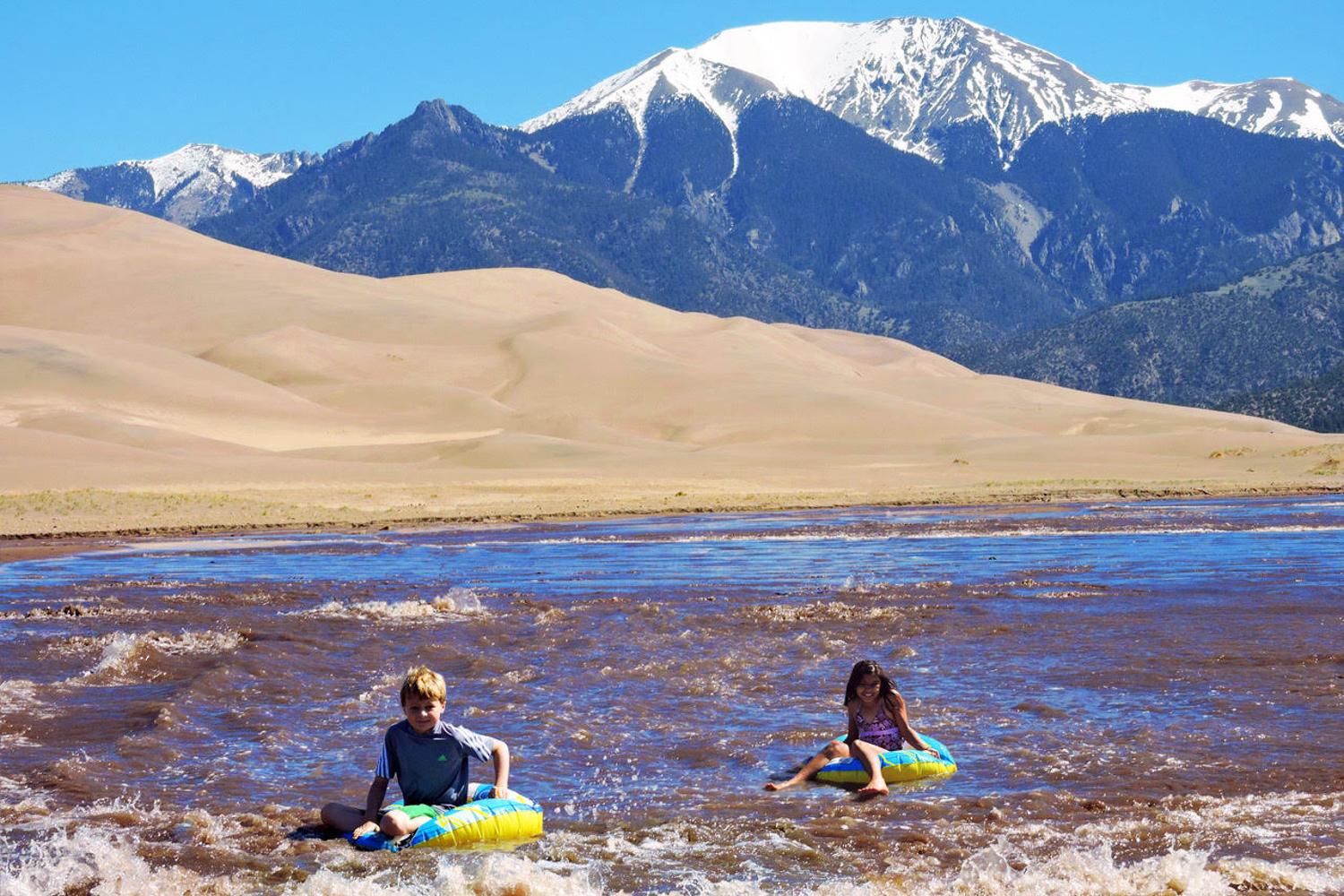 Two children are riding inflatable toys in flowing water with sand dunes and snowcapped mountains behind them