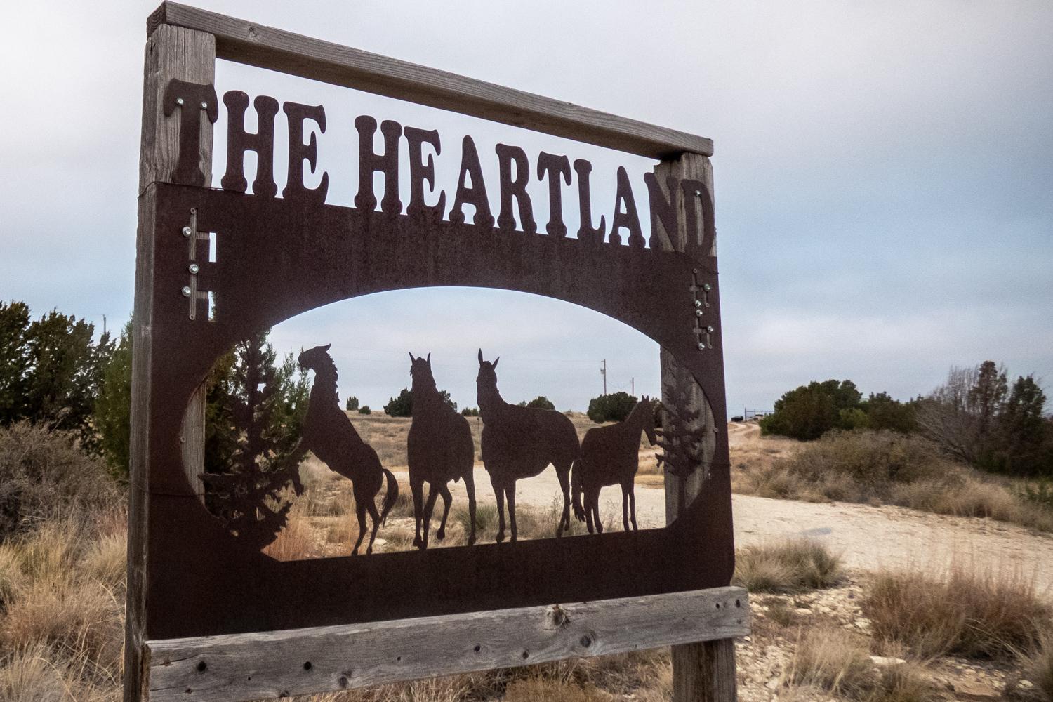 sign with a silhouette of horses cut from metal and the words "The Heartland" A dirt road runs past it