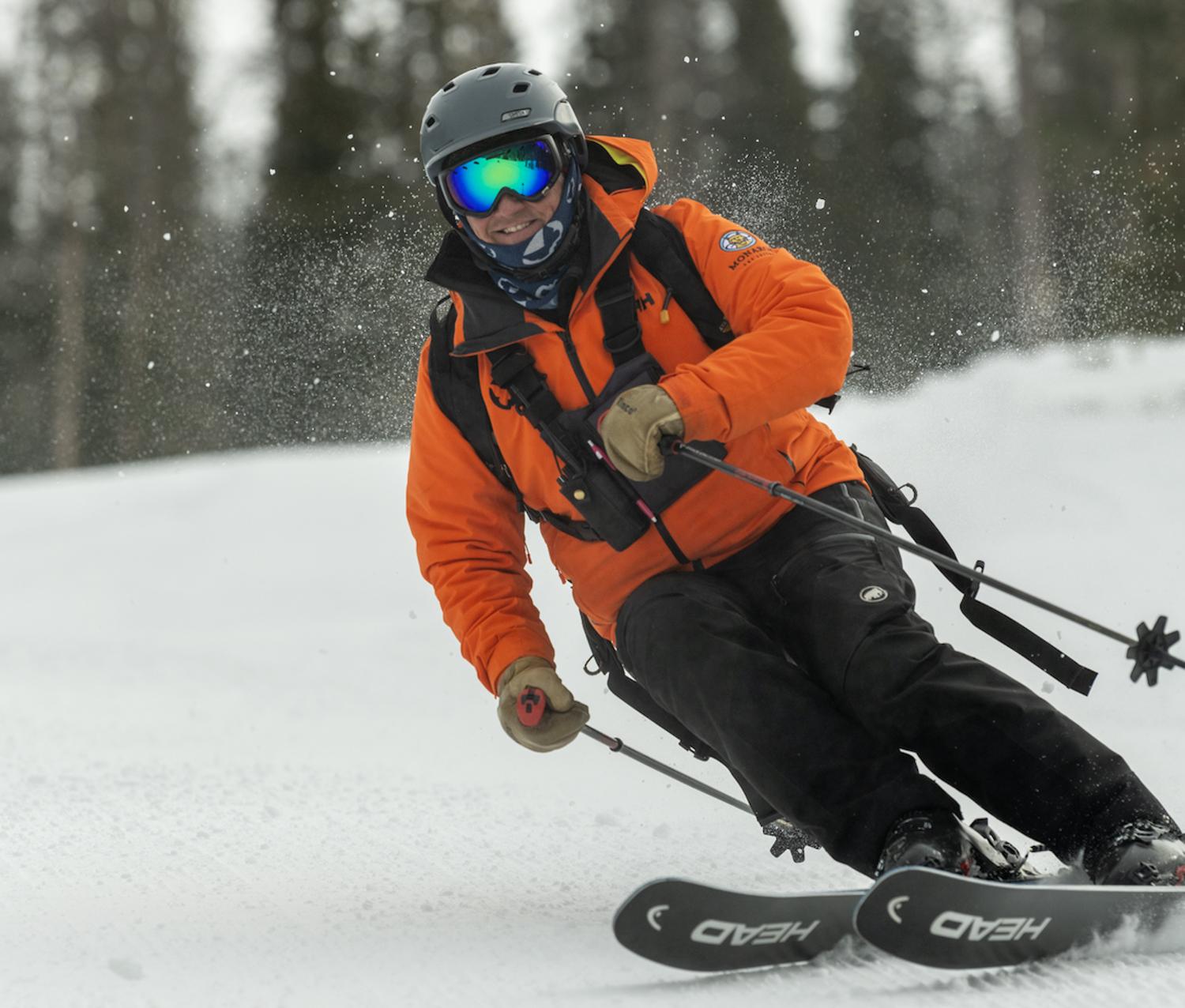 A smiling person wearing an orange jacket and a backpack is skiing down a snowy slope.