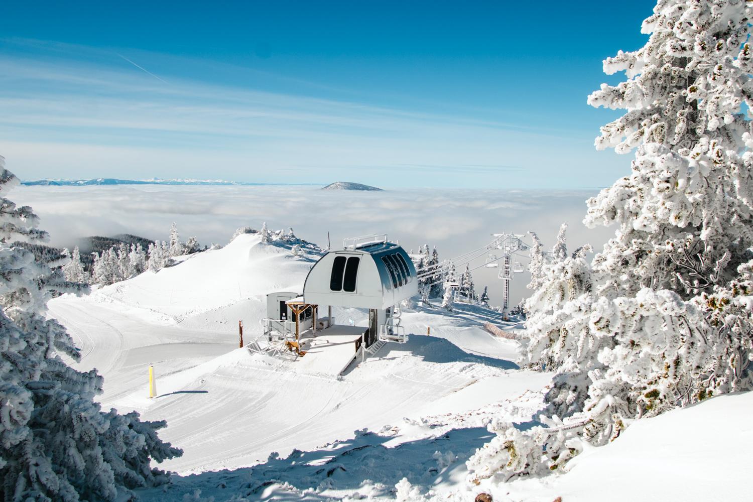 Snow covered mountain top with chairlift terminal above the clouds with blue sky, scene is framed by snow covered trees