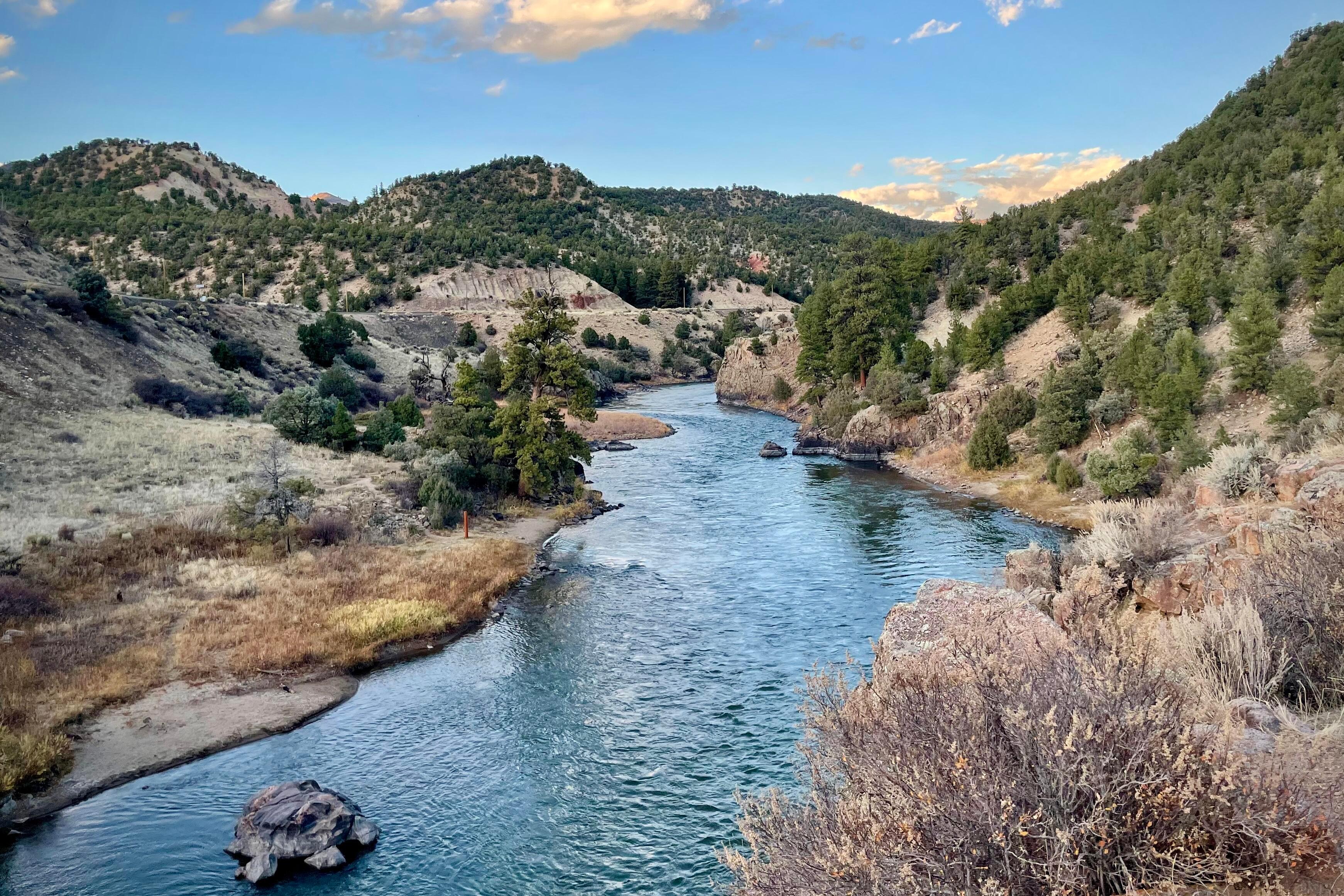 A shiny blue River flows through a mountainous area dotted with green trees. Dry willows and grass line the banks.