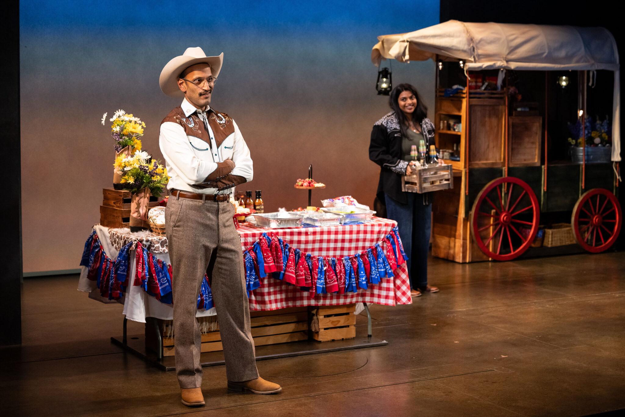 An Indian man stands wearing cowboy garb while his daughter looks on.
