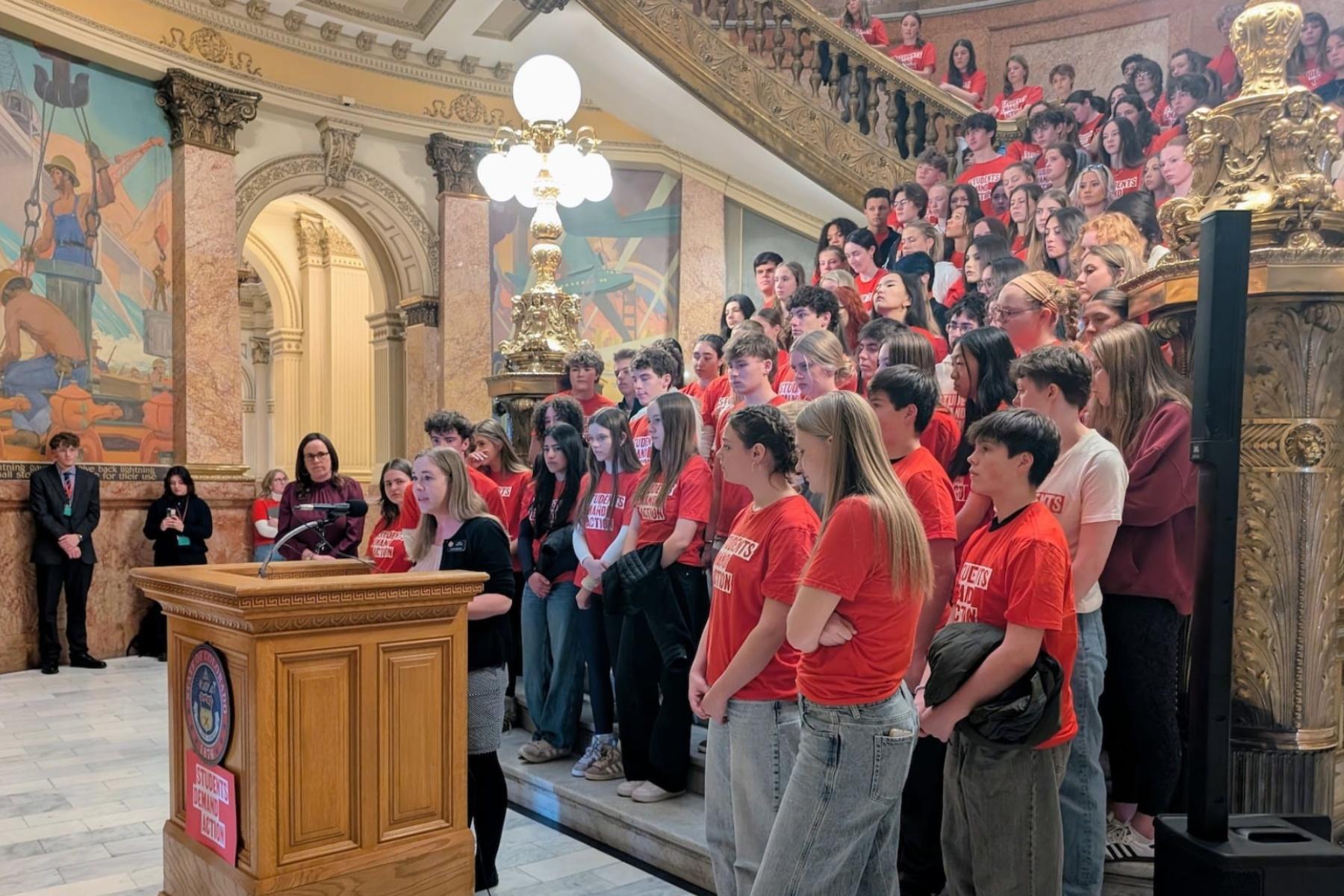 Students in red t-shirts stand on a large stair case and watch another student speaking at a podium.
