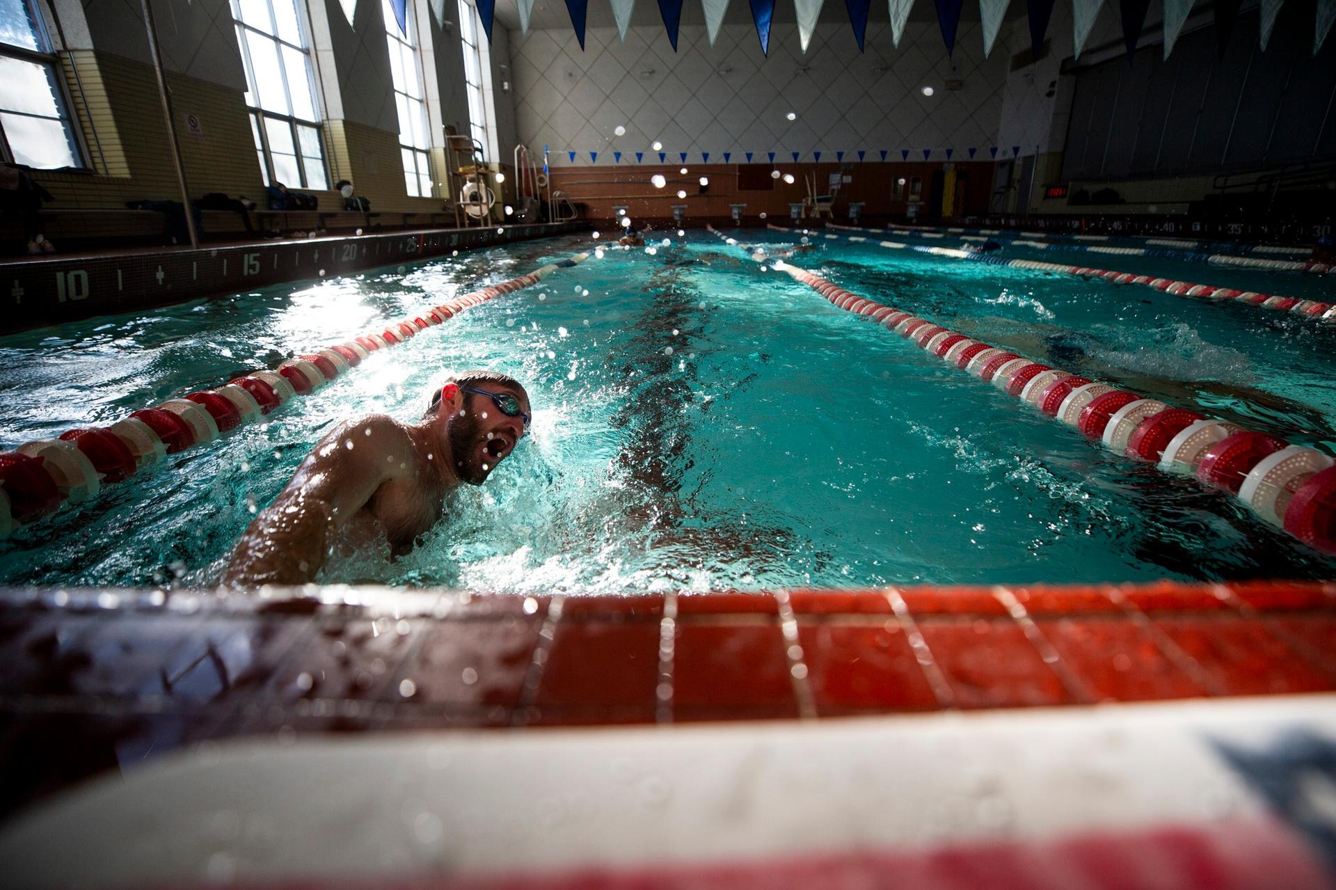 Water droplets are suspended in the foreground as a man wearing swim goggles launches off the wall of an azure pool stretching into the background.