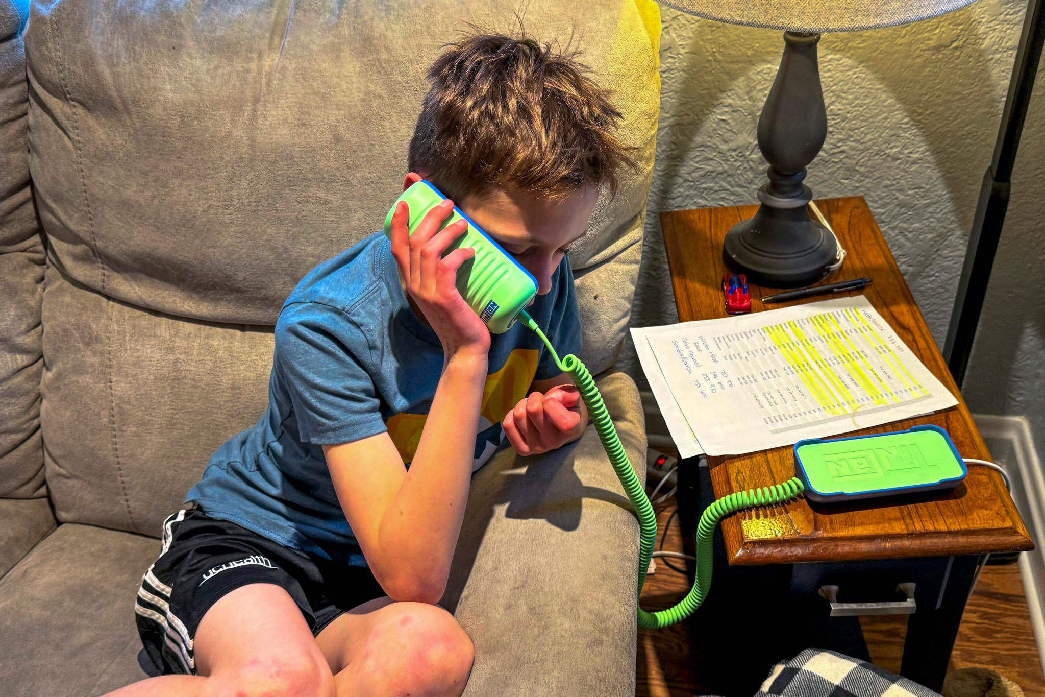 A kid in a t-shirt and shorts sits on a couch, leaning on the arm rest as he talks into a green land-line phone.