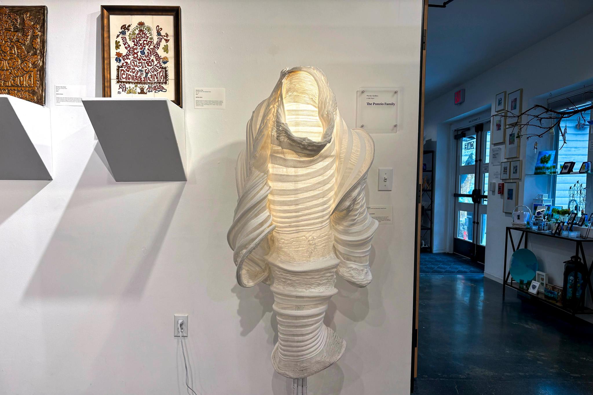 An object made of glowing, frilly fabric evokes the shape of a person — or a ghost — as it hangs on a white wall.